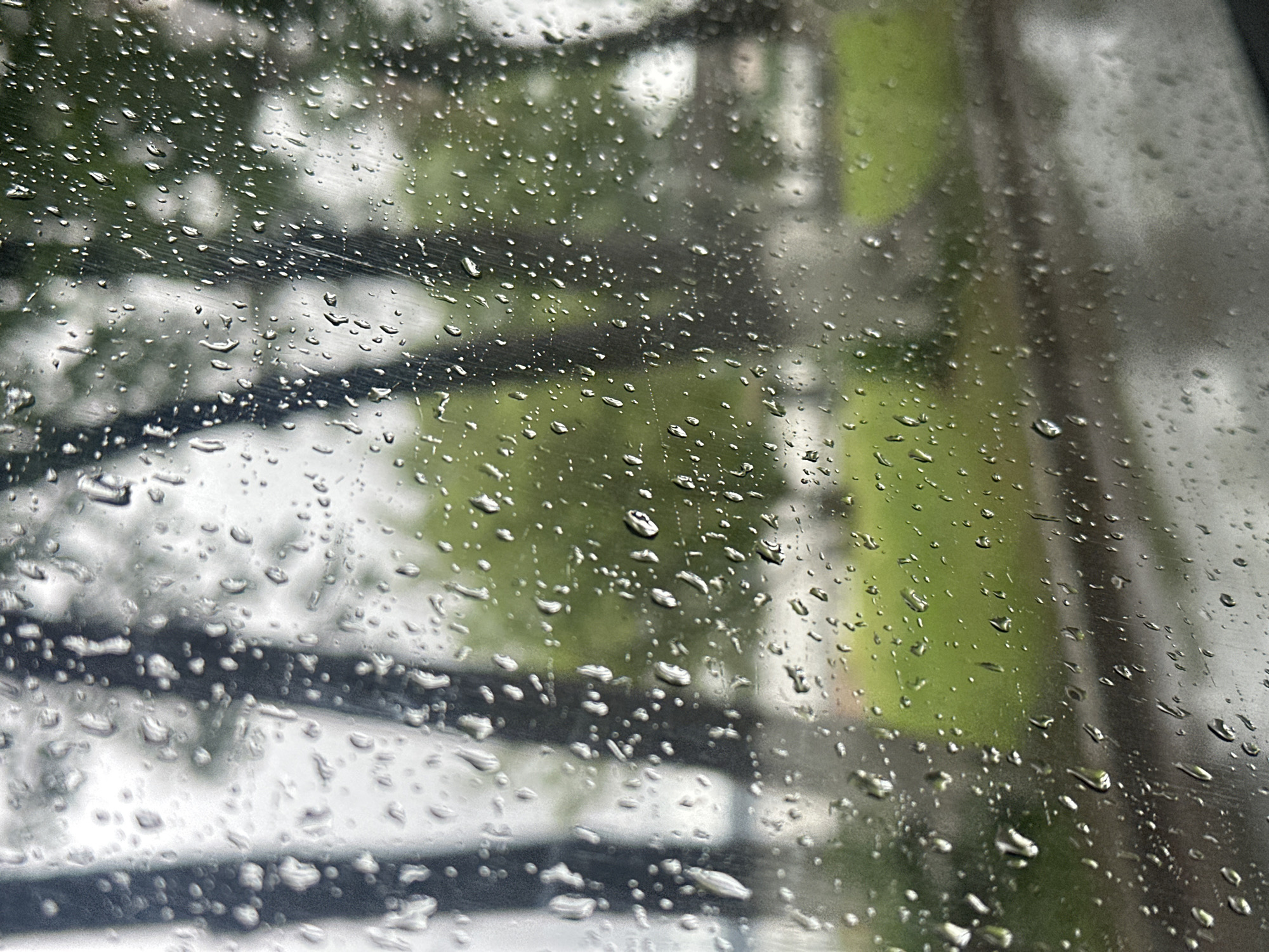 The image depicts a rainy day scene viewed through a window, likely in Votkinsk, Russia, though without external location cues, direct confirmation is impossible. Raindrops obscure the view, blurring the background into indistinct shapes and colors. A black, metallic structure is visible through the rain, likely a railing or fence, creating a geometric pattern contrasting with the organic shapes of the raindrops. Behind the structure are blurry green patches suggesting foliage, and some indistinct forms that could be buildings. The light is muted, consistent with overcast conditions. The focus is on the raindrops on the glass surface.