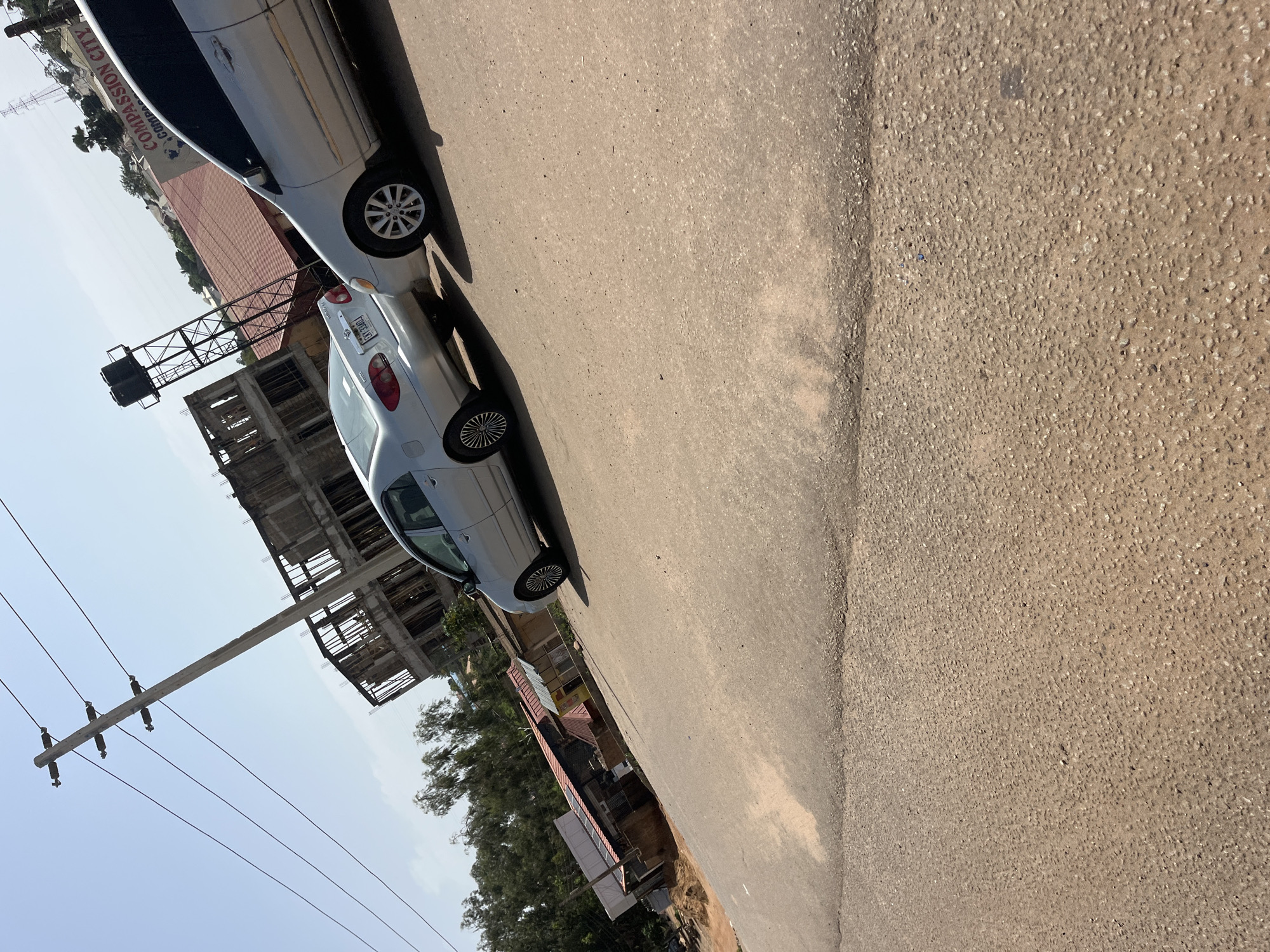 The image shows a street scene with two silver cars parked on a paved road. In the background, there is an unfinished concrete building under construction, a water tower, and various structures with red roofs. A utility pole with wires is visible on the left side of the image. A sign with "COMPASSION CITY" written on it is also present. The sky is clear and blue. The image is taken from a slightly low angle, looking up towards the buildings and sky. The ground is a mixture of asphalt and dirt.