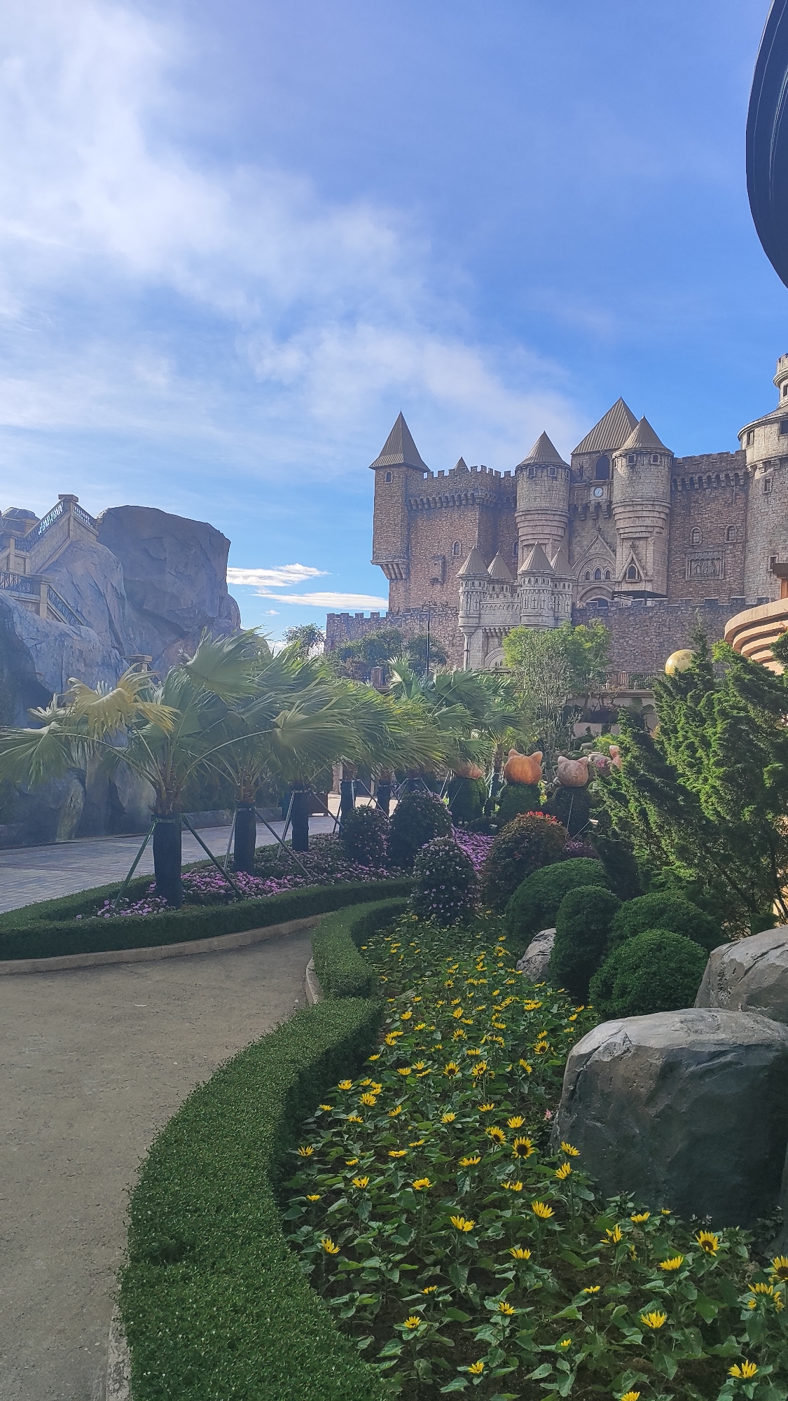 The image depicts a fantastical castle, reminiscent of European medieval architecture, situated in a theme park setting. The castle is constructed of stone and features multiple turrets, towers, and battlements, with a clock visible on one of the central towers. In the foreground, a landscaped garden with manicured hedges, palm trees, and blooming yellow flowers curves along a pathway. The scene is bathed in daylight under a blue sky with scattered clouds. This location is Sun World Ba Na Hills in Đà Nẵng, Vietnam, a popular tourist attraction known for its French village and fantasy-themed attractions, including this imposing castle structure. There are no people or animals visible in the image. The overall impression is one of a meticulously designed recreational area, evoking a sense of fantasy and escapism.