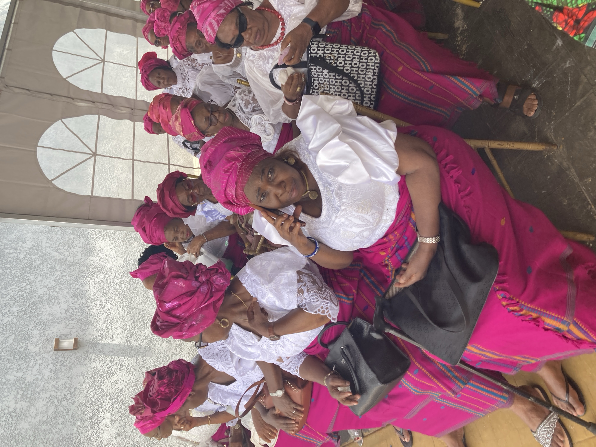 A group of women are seated together in what appears to be a tent or covered space. They are uniformly dressed with pink headwraps and white tops with pink skirts. Many are wearing jewelry, including necklaces and watches. Some are holding purses. The seating appears to be made of bamboo. The background is mixed, with tent walls and what may be a building. The location is Nnenu, Nigeria.