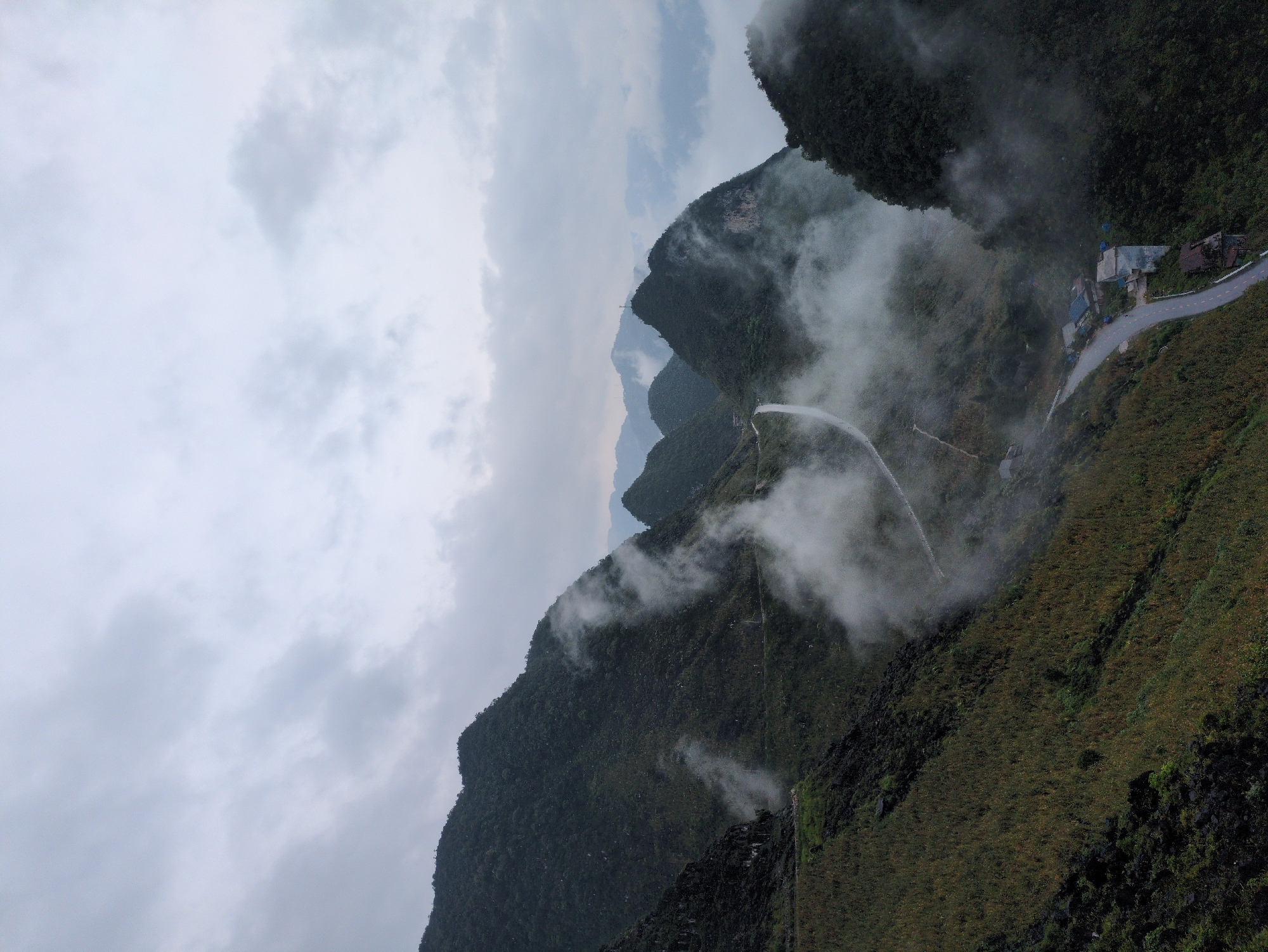 This image captures a misty, overcast scene of the Ma Pi Leng Pass in Dong Van Commune, Vietnam. The pass is characterized by its dramatic, mountainous landscape, with steep, verdant slopes rising sharply. A winding road snakes through the valley, appearing as a thin grey ribbon against the dark green of the mountainside. Wisps of white fog drift through the valleys and cling to the mountains, obscuring parts of the landscape and creating a sense of depth and mystery. In the lower right of the image, a few small, clustered buildings are visible, nestled along the roadside, suggesting a small settlement. The overall atmosphere is one of rugged, natural beauty, amplified by the atmospheric conditions.