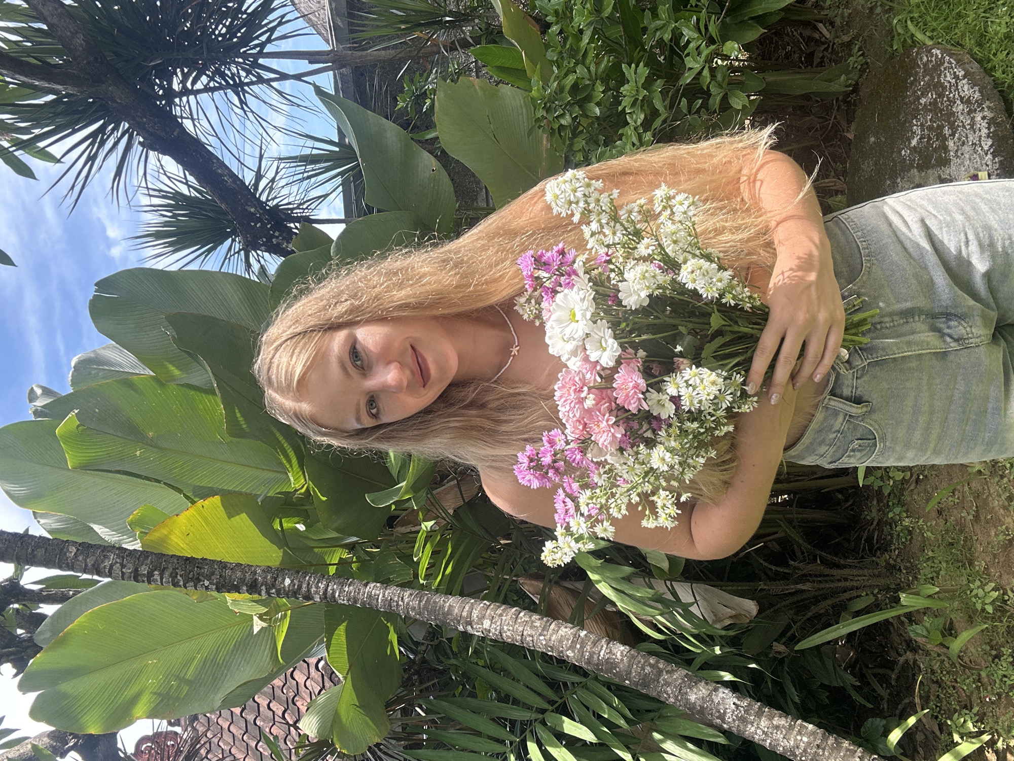 The image captures a young woman with long, wavy blonde hair, gently smiling and looking towards the viewer, holding a large bouquet of flowers outdoors in a tropical setting. Her head is slightly tilted, and she wears a delicate white beaded necklace with a small star pendant.
The bouquet she holds is vibrant, comprising a mix of white, pink, and purple small flowers. Her left hand, adorned with light-colored nail polish, supports the flowers, with sunlight casting dappled shadows on her arm. She appears to be wearing light-colored denim, possibly shorts or overalls, and a light-colored top, revealing her shoulders.
The background is filled with lush tropical foliage, including large, broad green leaves and the textured trunks and fronds of palm trees reaching towards a clear blue sky with scattered white clouds. The strong sunlight indicates it is daytime, likely midday or early afternoon. Hints of a tiled roof structure are visible in the background, suggesting a building or garden feature. The overall scene conveys a serene and natural atmosphere, aligning with the tropical environment of Kuta Selatan, Indonesia. There is no visible text in the image.