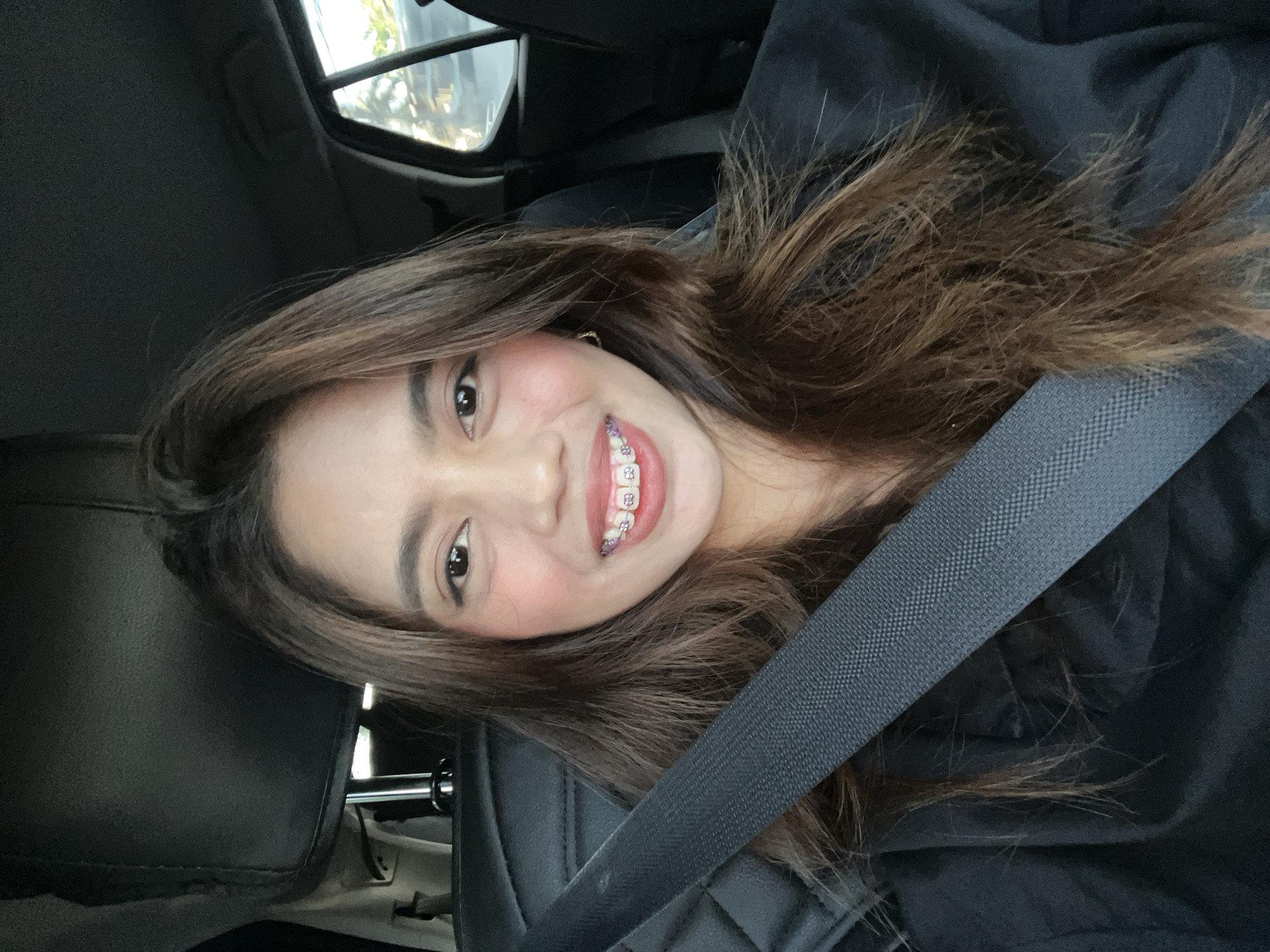 A young adult female, consistent with Southeast Asian ethnicity, is seated in the front passenger seat of a vehicle in Davao City, Philippines. She has long, dark brown hair with lighter brown highlights, parted on her left, and dark eyes. Her face shows visible blush on the cheeks, and she wears small gold hoop earrings. She is smiling widely, displaying clear teeth fitted with purple orthodontic braces on both upper and lower arches. She is wearing a dark, likely black, long-sleeved garment. A black fabric seatbelt, featuring a checkered texture, extends diagonally across her chest from her right shoulder to her left hip. The car's interior is dark, with a black headrest visible behind her head and a black textured seat. Through the rear passenger window on the right side of the frame, a sunlit outdoor environment is visible, including green foliage resembling palm trees and a dark-colored vehicle in the background, indicating daytime conditions.