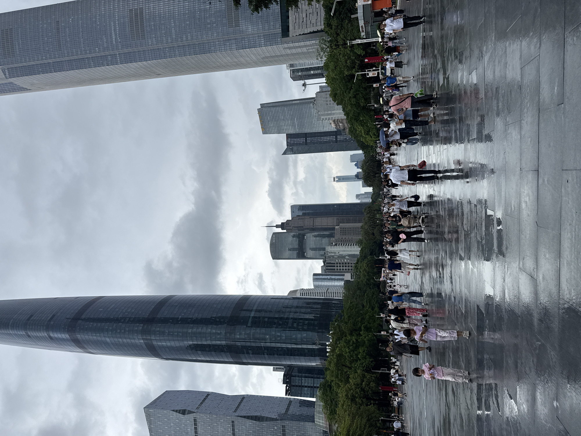In Tianhe District, China, a wide, wet plaza is filled with pedestrians walking under an overcast sky. The plaza's dark, reflective stone surface is wet, creating mirrored images of the people and surrounding architecture. Towering skyscrapers dominate the background, including the distinct cylindrical design of the Guangzhou IFC and the angular facades of other modern buildings. Lush green trees line the edge of the plaza, separating it from the bustling urban landscape. People of all ages are scattered across the plaza, some walking in groups, others alone, with a few children visible near the foreground. The atmosphere appears to be a normal day in a busy city, with people going about their business despite the damp conditions.