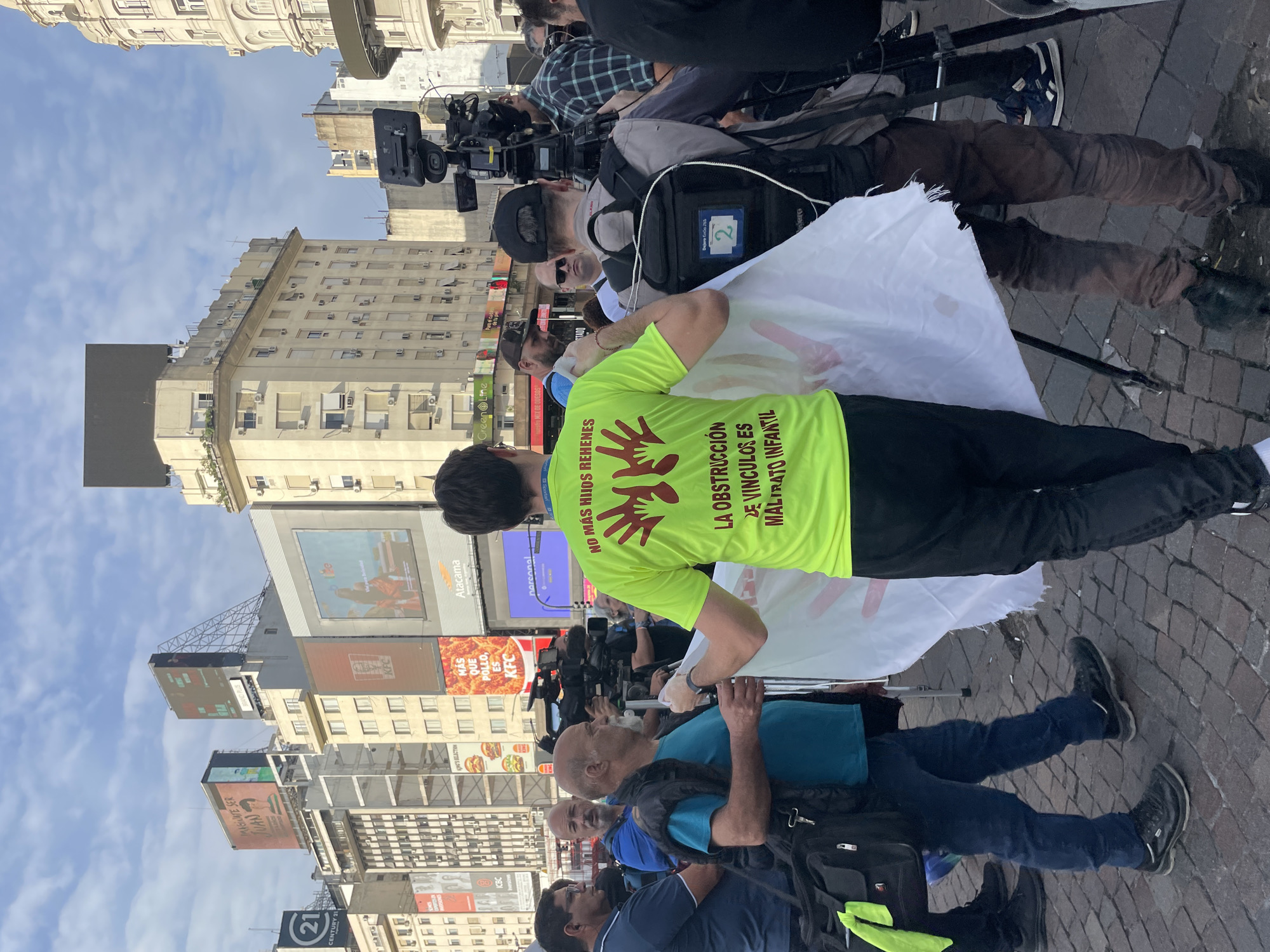The image captures a protest in a bustling urban area, likely Buenos Aires, Argentina, indicated by the architectural style and billboards. A group of individuals are gathered on a paved street, with a man in a bright green t-shirt as a focal point. His t-shirt has a printed message in Spanish, along with handprint graphics. Around him, people hold a white banner. Multiple news cameras and reporters are present, suggesting the protest is attracting media attention. The background features a mix of buildings, including skyscrapers with digital billboards displaying various advertisements. The overall environment is busy and suggests a central location within a city.