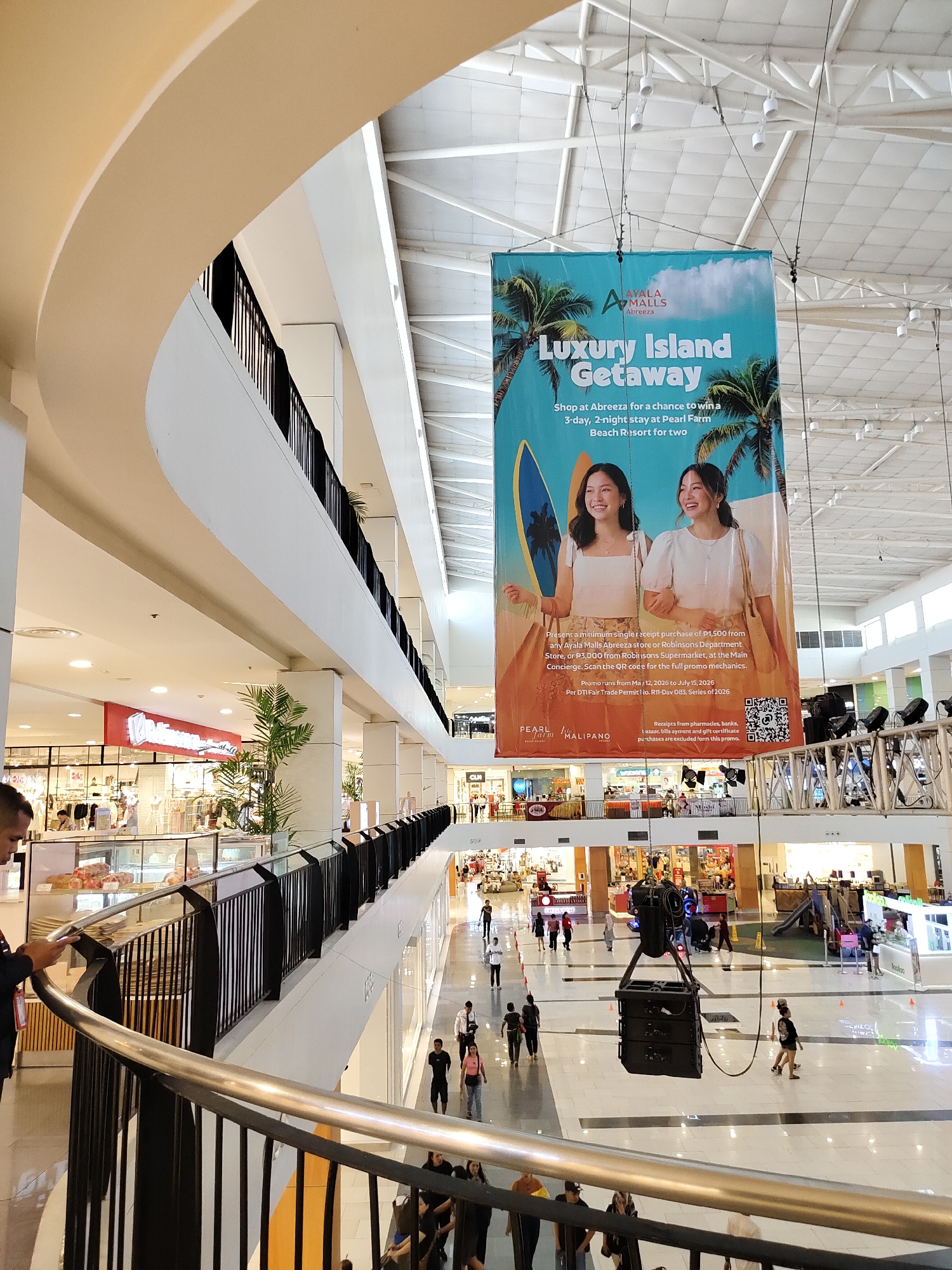 The image shows the interior of a mall in Davao City, Philippines. A large banner promoting a "Luxury Island Getaway" is suspended from the ceiling. The banner features images of two young women, palm trees, and surfboards, and advertises a chance to win a three-day, two-night stay at Pearl Farm Beach Resort. Text on the banner indicates that this promotion is at Ayala Malls Abreeza. The mall appears to be multi-level, with escalators and railings visible. Several people are seen walking around the mall, browsing shops, and interacting. The environment is brightly lit with a high ceiling. Various shops and kiosks are present, as well as a children's play area in the background. A large speaker system is also suspended from the ceiling in the foreground.