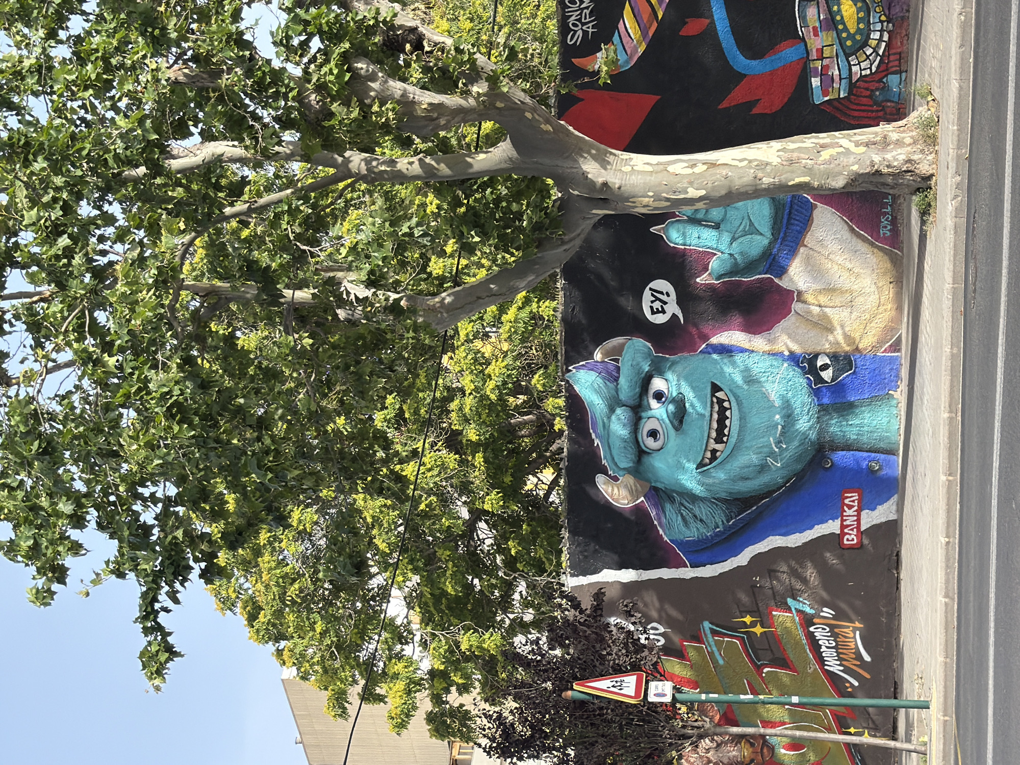 This is a full, eye-level, outdoor shot of a graffiti mural on a wall in a city. The mural features a large, turquoise, furry monster character with big eyes and horns, reminiscent of Sulley from Monsters, Inc. The monster is depicted from the chest up, wearing a blue jacket with yellow accents. A speech bubble above its head says "Ey!". To the left of the monster, there is graffiti with the name "Moreno Mural" written in stylized script, along with red and yellow spray paint. Further to the left, a street sign with a red border and a white background depicts two figures walking, with smaller text below. A tree with a thick, light-colored trunk and branches covered in green leaves stands in front of the mural, partially obscuring the graffiti on the right side of the image. Behind the mural and tree, there are green trees and a building with a beige facade. The mural is on a dark wall, with various colorful elements and abstract designs visible. The foreground shows a paved sidewalk and a street with road markings. The sky is clear and blue. The scene appears to be in a public area, possibly a street or alleyway.