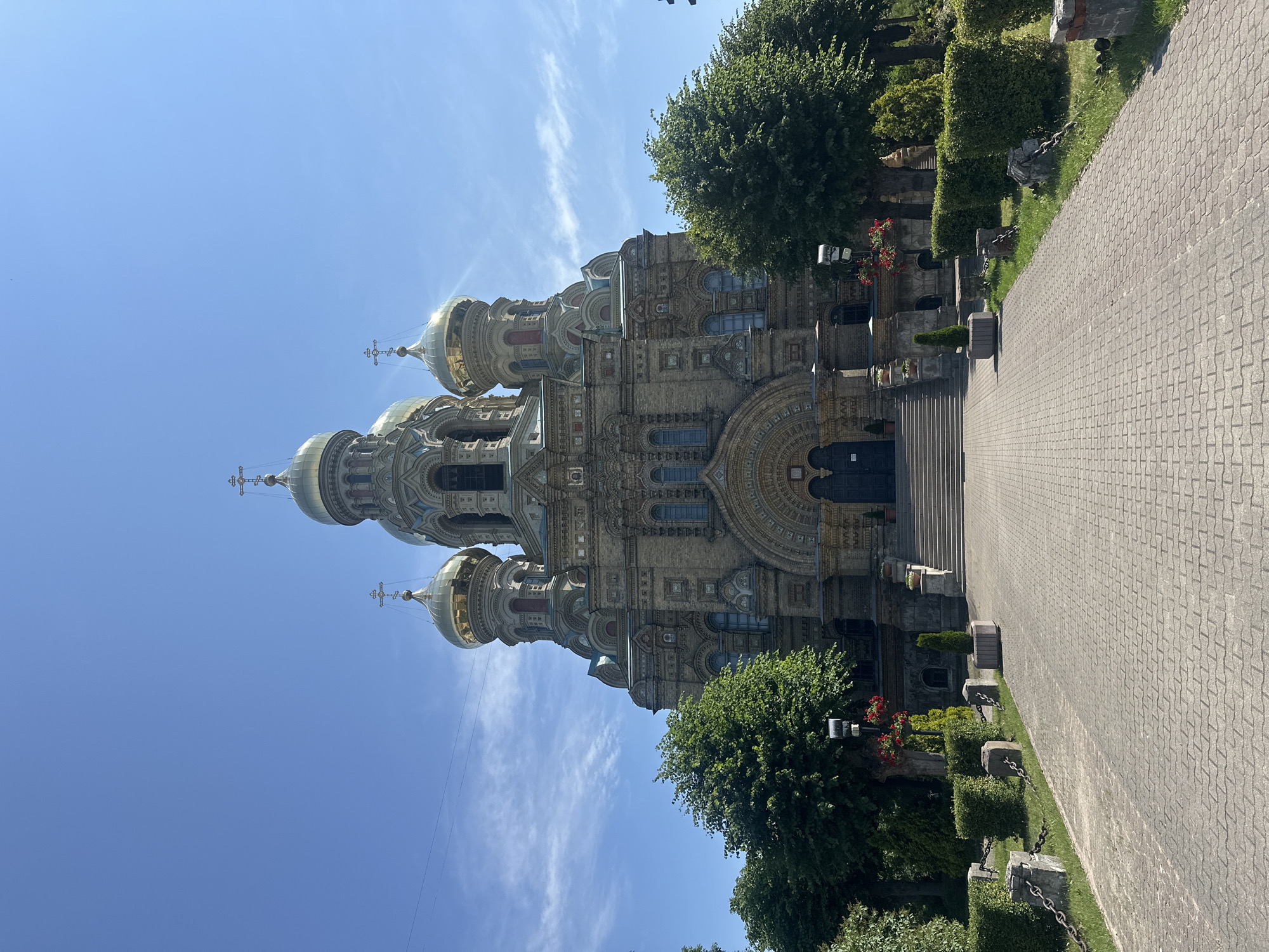 The image shows the Church of St. Nicholas Naval Cathedral in Liepaja, Latvia, under a clear blue sky with scattered clouds. The large, ornate cathedral dominates the center of the frame, featuring multiple golden domes topped with crosses. The facade is constructed of light-colored brick with intricate architectural details and arched windows.
In front of the cathedral, a paved walkway made of interlocking bricks leads up to a wide set of stairs. The walkway is flanked by manicured green hedges and trees. Along the left side of the walkway, there are low stone pillars connected by chains, interspersed with grass and more hedges. A few potted plants and decorative flower arrangements are visible near the entrance and on the stairs. The scene appears calm and the cathedral is the sole focus. There are no people visible in the image. The lighting suggests it is daytime, likely mid-morning or afternoon, given the bright sunlight.