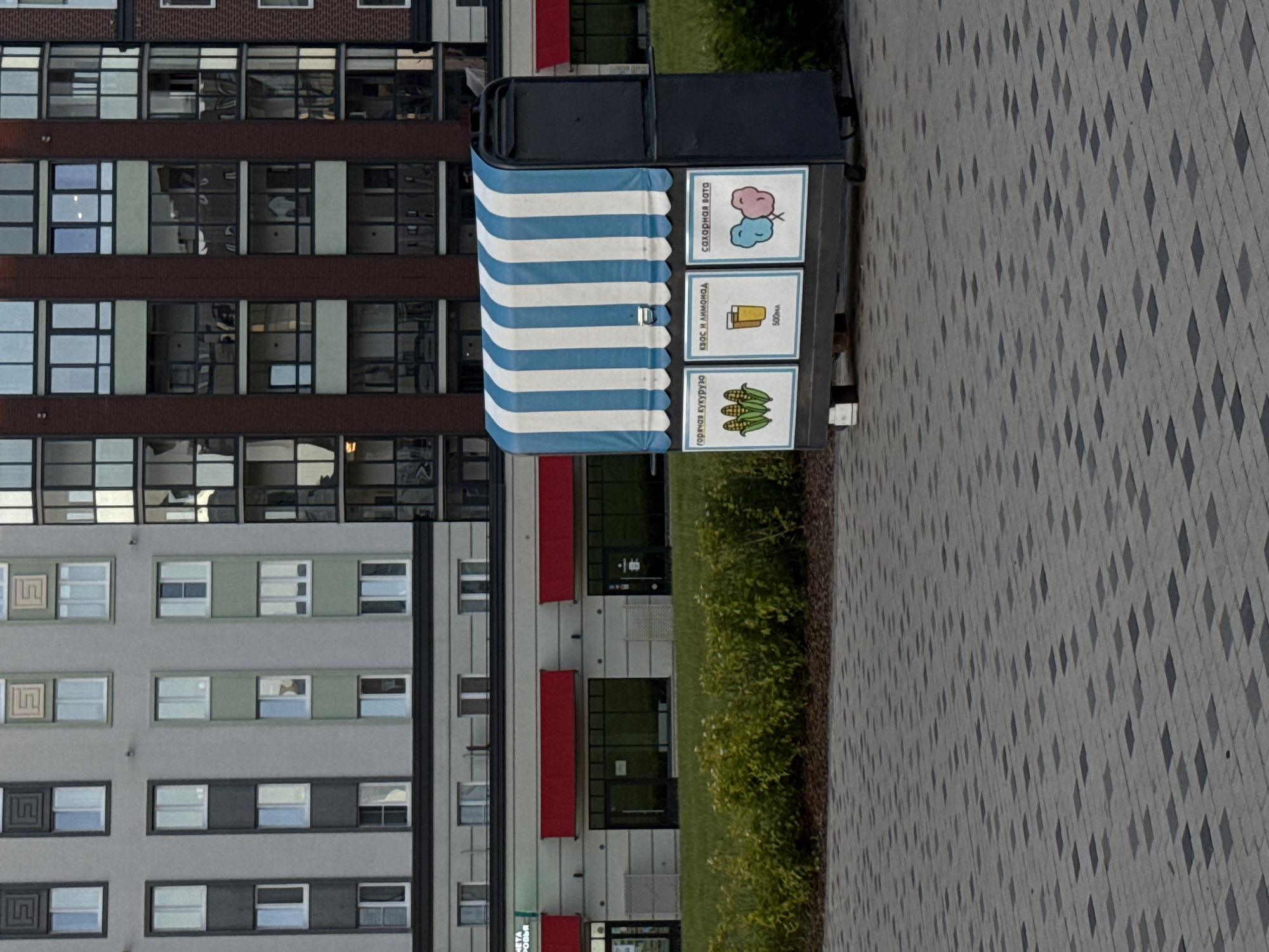 This is a medium shot, eye-level, outdoor, daytime photograph taken in Izhevsk, Russia. The foreground features a paved walkway with a diamond pattern of light and dark grey paving stones. In the middle ground, there is a small, dark grey food cart with a blue and white striped awning. The cart displays three signs: one with an illustration of corn labeled "ГОRЯЧАЯ КУКУРУЗА" (hot corn), another with an illustration of a glass of juice and the text "КВАС И ЛИМОНАД 500мл" (kvass and lemonade 500ml), and a third with an illustration of two cotton candy puffs labeled "САХАРНАЯ ВАТА" (cotton candy). Behind the food cart is a strip of green grass and low-lying bushes. In the background, a modern multi-story building with a light grey facade and rows of windows is visible on the left. To the right of the grey building is another building with a dark red brick facade and dark-paned windows, also with multiple stories. The sky is not visible in the frame.