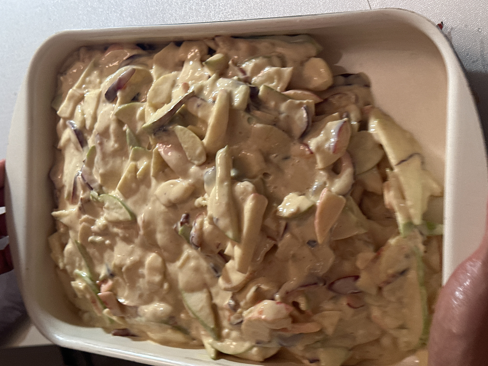 An overhead shot shows sliced apples mixed with a creamy, light brown sauce in a white ceramic baking dish. The dish is filled almost to the brim, with some of the mixture spilling over the side onto the white countertop. The apples appear to be a mix of green and red varieties, sliced thinly and unevenly. The sauce is thick and covers most of the apple slices. The background is blurred, but it appears to be a domestic setting. There is no indication of the city of Nevinnomyssk, Russia.