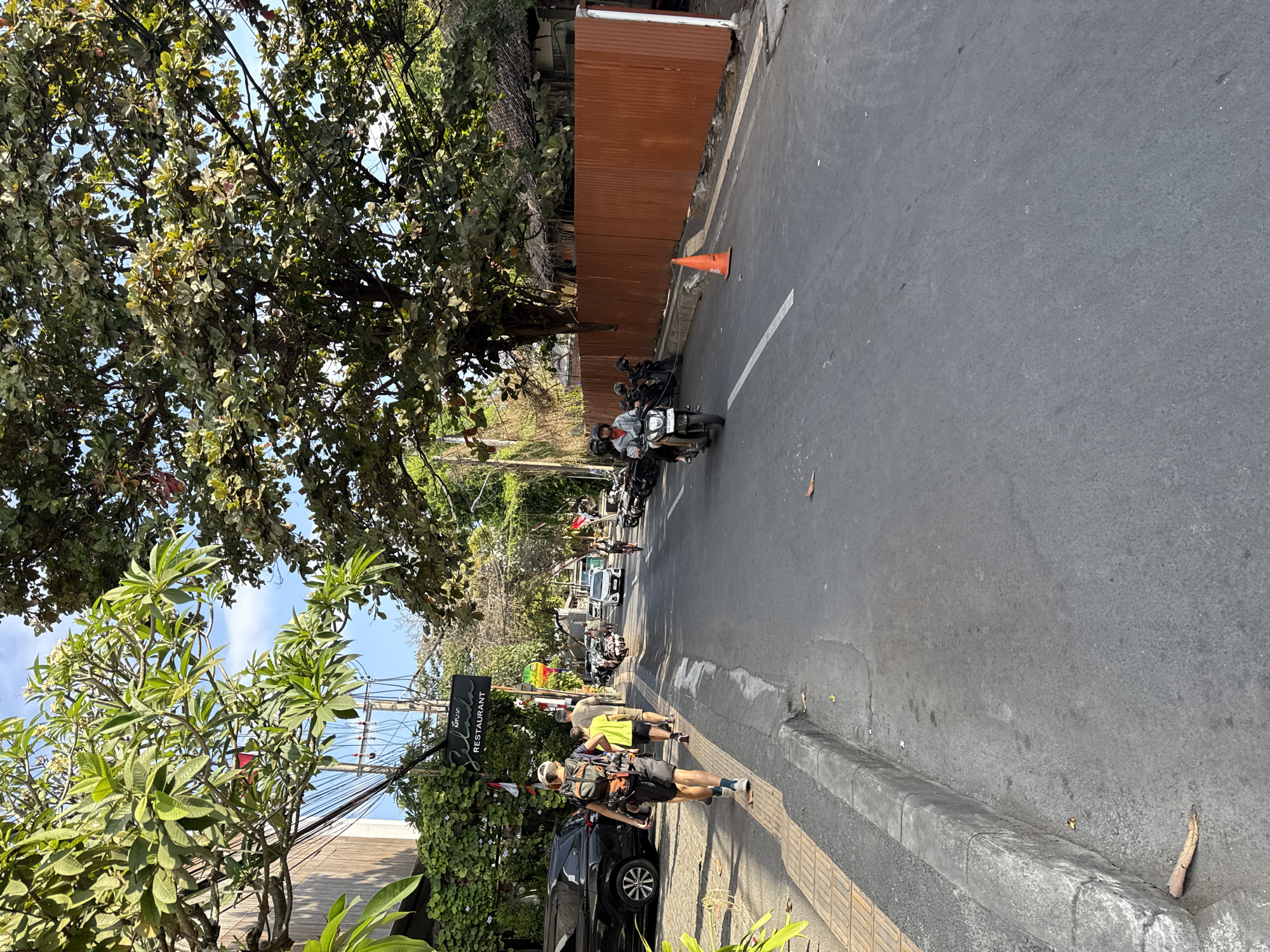 The image depicts a street scene in Denpasar, Indonesia, likely during daylight hours. In the foreground, a man with a backpack walks away from the camera on a sidewalk. To his left, a dark-colored SUV is parked along the side of the road. Further down the street, several motorcycles are parked on both sides of the asphalt road. Two people are riding a motorcycle, with the driver wearing a helmet and the passenger behind them. An orange traffic cone is positioned on the road near a rusty metal fence. Trees with lush green foliage line the street, casting shadows. A sign for "Selasa RESTAURANT" is visible on the left side of the road. The overall environment suggests a bustling urban or suburban setting.
