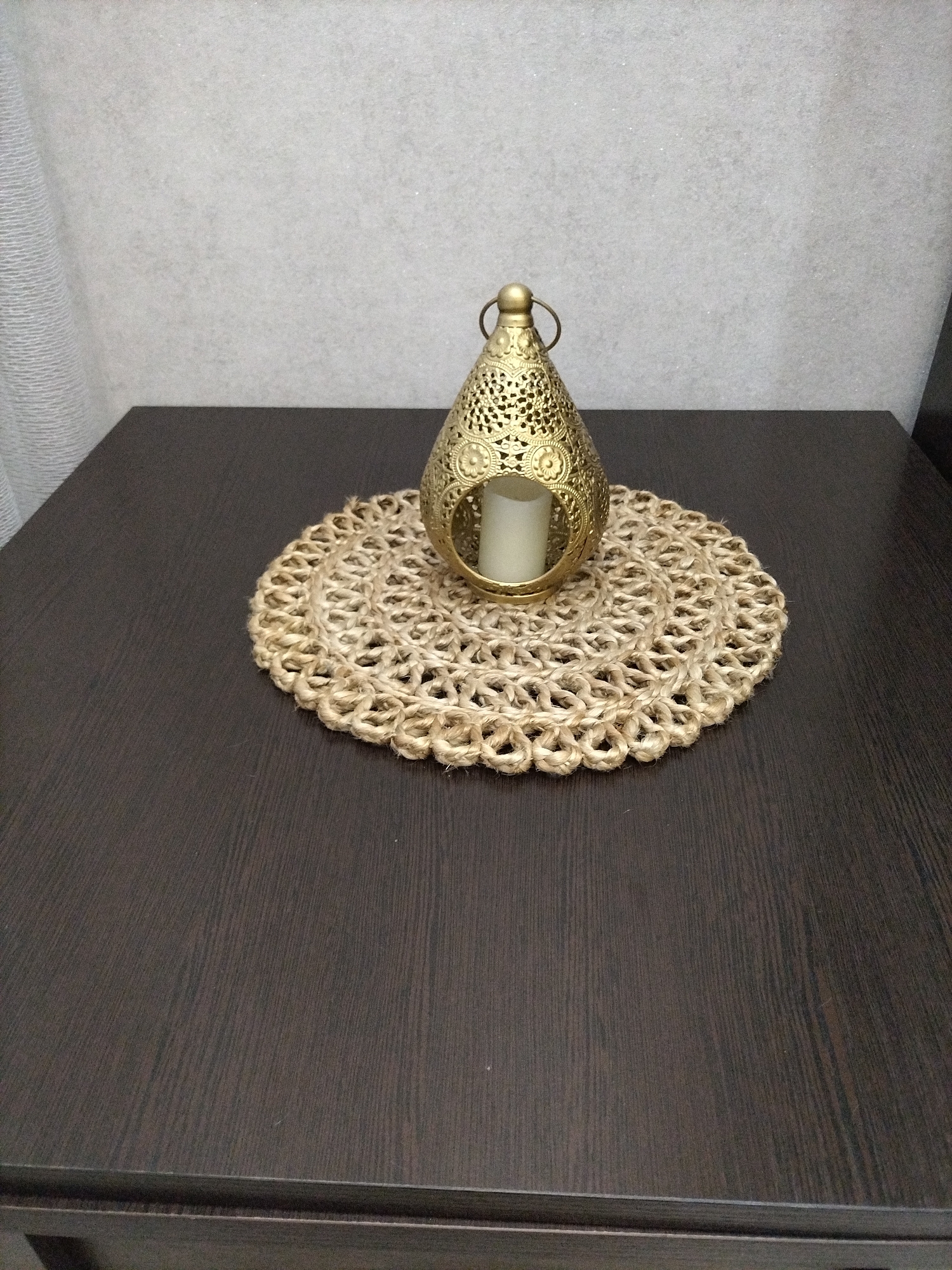 The image shows a close-up, eye-level shot of a decorative brass lantern with a candle inside, placed on a jute placemat. The placemat is circular and has a woven, textured pattern. The lantern itself is ornate, featuring cut-out designs and a small handle at the top. It is positioned slightly off-center on the placemat, which rests on a dark, wooden surface, likely a table. The background consists of a textured, light-colored wall, with a hint of a white curtain on the left side of the frame. The lighting appears to be indoor, with soft illumination that highlights the metallic sheen of the lantern and the natural fibers of the placemat. No people are visible in the image, and there are no discernible signs of text. The overall impression is one of home decor, suggesting a quiet and perhaps cozy atmosphere. The context provided places this scene in Nizhnyaya Makhtama, Russia.