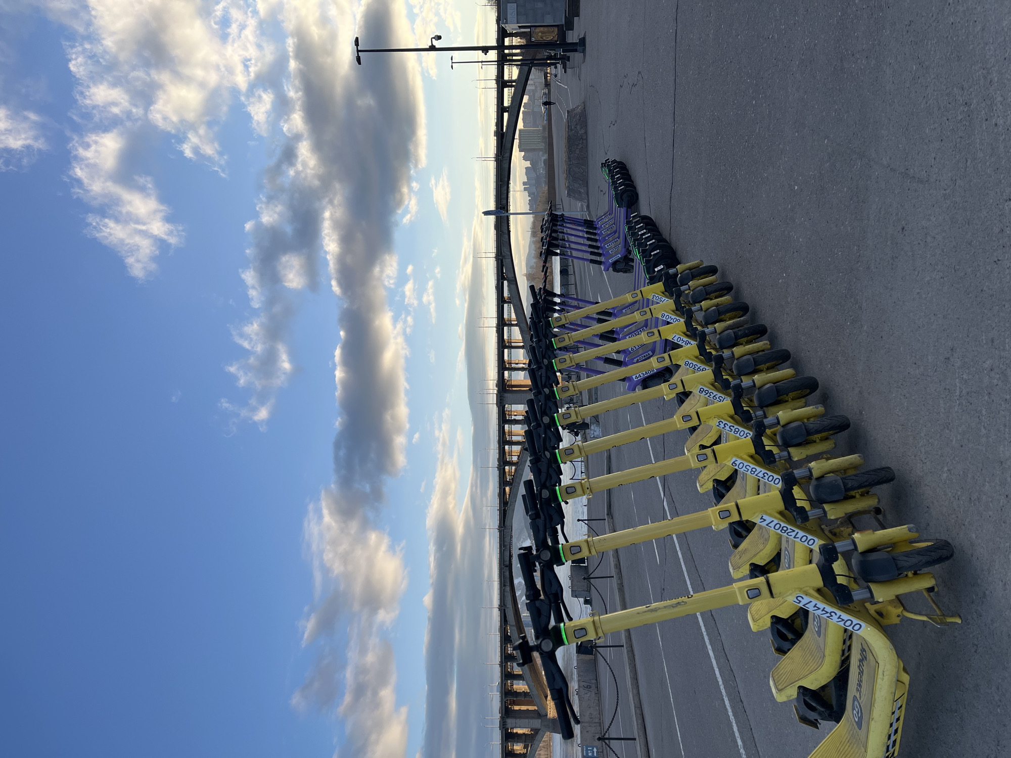The image, taken in Krasnoyarsk, Russia, captures a serene urban scene during what appears to be the golden hour, either late afternoon or early evening. The sky is partly cloudy, with white and grey clouds illuminated by warm sunlight, contrasting with clear blue patches.
In the foreground, multiple rows of rental electric scooters are neatly parked on an asphalt surface. The prominent yellow scooters feature "Яндекс Go" branding, clearly visible on their front plates, along with various identification numbers such as "00434475", "00128074", "00378509", "308533", "159368", "09308", "48407", "00408", "1503", "6070", "442984", "443408", "507466", "61", and "403215". Further back, another set of electric scooters with purple bodies and black accents are also lined up.
The setting includes a wide, multi-span bridge in the mid-ground, likely crossing a large river such as the Yenisei. Beyond the bridge, a distant cityscape with numerous buildings is visible, bathed in the soft light of the low sun. Streetlights and utility poles punctuate the scene, suggesting a well-developed urban environment. There are no people visible, giving the impression of a quiet moment in a busy city. The overall atmosphere is tranquil, highlighting urban infrastructure and modern transportation options under a dramatic sky.