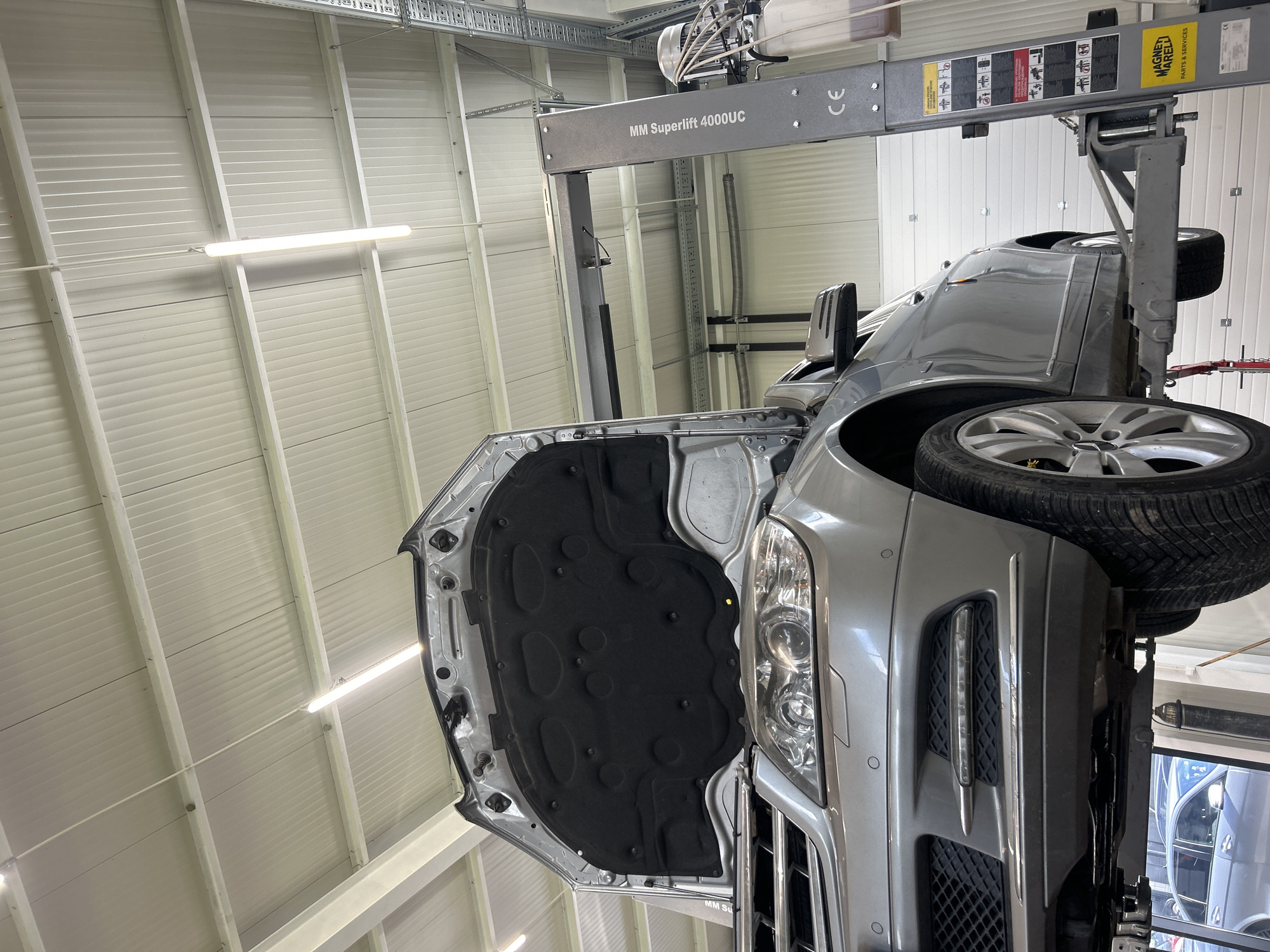 The image shows a silver-colored car, specifically the front portion including the hood, headlight, and front passenger side tire, raised in the air by a "MM Superlift 4000UC" vehicle lift inside what appears to be a car repair shop or garage. The hood is open, revealing a black underhood thermal insulation liner. The shop has a white, corrugated metal ceiling with fluorescent lights. The exact location in the city of Tsaratsovo, Bulgaria cannot be confirmed from the visual information provided.