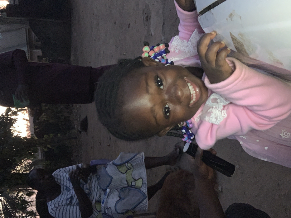 A smiling, dark-skinned child in a pink, lace-trimmed dress looks directly at the camera. Colorful beaded decorations adorn her hair. Behind her, a person in a dark purple garment stands near a setting sun. Another individual, seated and wearing a light-colored patterned dress, is also visible. In the foreground, hands hold a black object, potentially a phone or remote. The scene appears to be outdoors, capturing a candid moment.