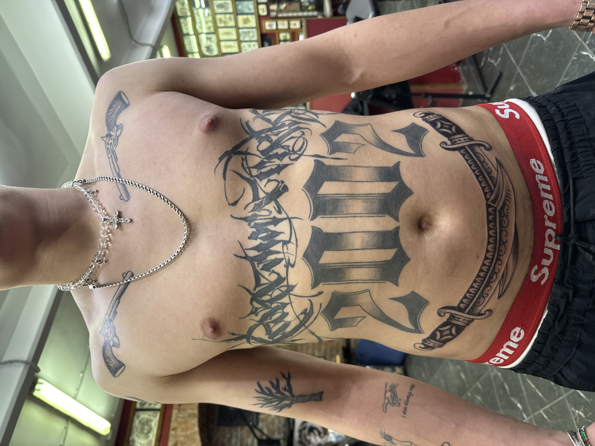 A person with numerous tattoos is depicted from the chest up. The tattoos include: two revolvers on either side of the chest; calligraphic designs with geometric shapes on the torso; a large design on the abdomen; a knife or dagger on the right side of the abdomen; and smaller tattoos on the left forearm, including a tree or plant. The person is wearing a necklace with a cross pendant, a watch, and Supreme-branded underwear. The background appears to be a tattoo parlor, possibly in Moscow, Russia.