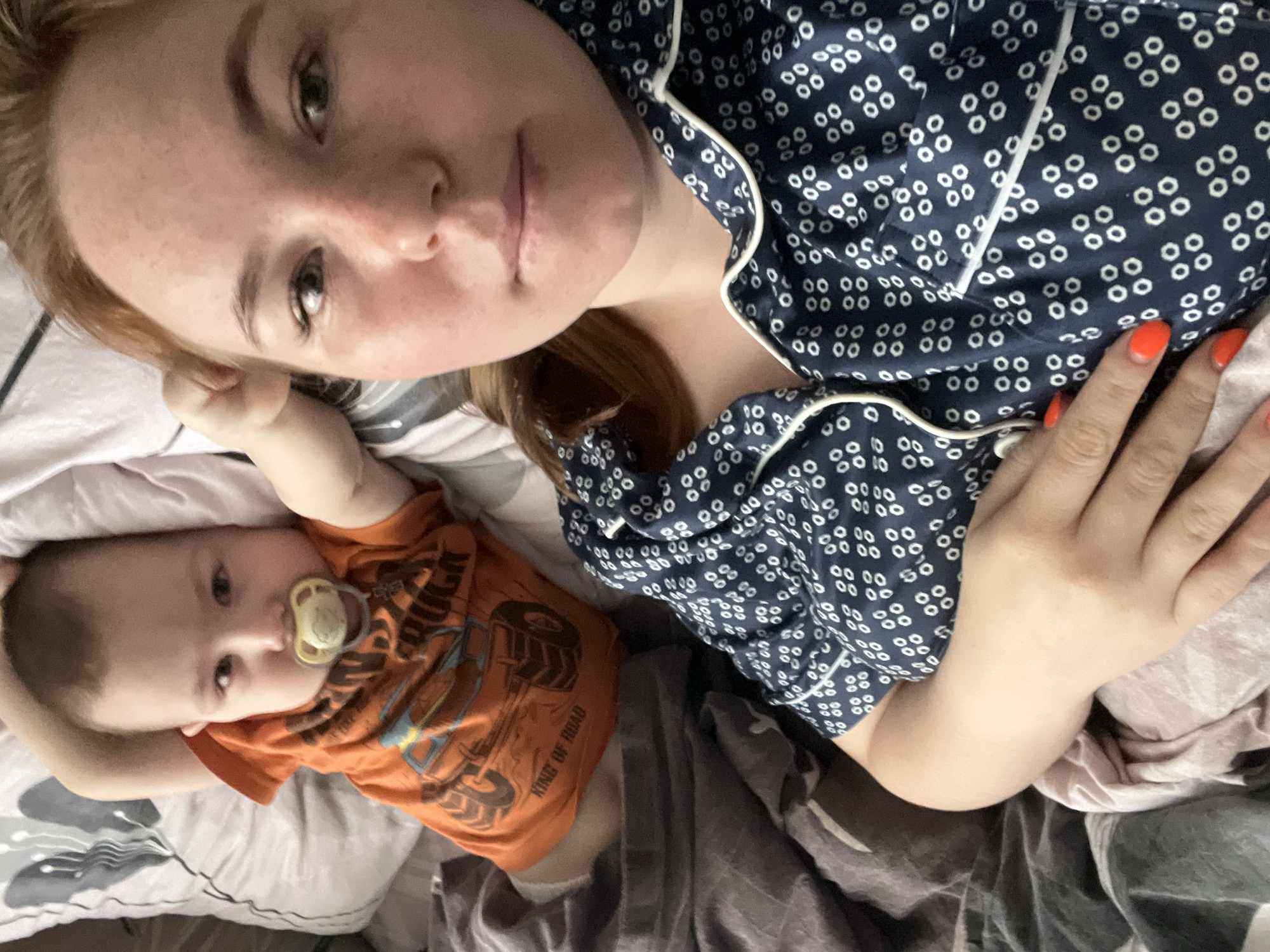 A person with fair skin and reddish-brown hair lies on their back in bed, wearing a navy blue pajama top with white trim and a pattern of white circles. Their right hand rests on their stomach. Lying next to them on the bed is a baby wearing an orange t-shirt with a monster truck graphic. The baby has a pacifier in their mouth. The background is a bed covered with a light-colored sheet and a gray blanket. There is no indication this photo was taken in Chaikovsky, Russia.