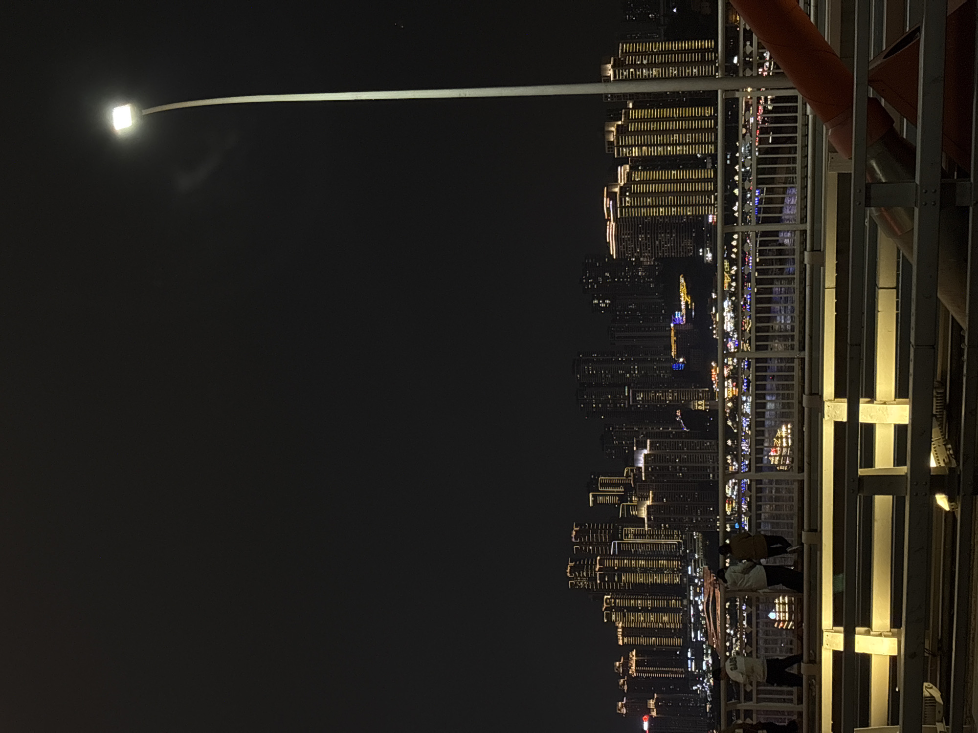 The image presents a nighttime panoramic view of a vibrant cityscape, likely from an elevated bridge or viewing platform in Yuzhong District, China. The scene is characterized by a stark contrast between the dark night sky and the dazzling array of artificial lights illuminating a dense urban environment.
On the left side of the frame, a modern street lamp extends prominently into the darkness, its bright square LED light serving as a primary light source in the foreground. Below and stretching across the horizontal mid-ground are numerous high-rise buildings, their countless windows glowing with predominantly warm yellow and white lights, creating intricate geometric patterns. Some buildings feature distinctive vertical strips of illumination, highlighting their architectural form. Further down and in the distance, a lower band of city lights includes hints of cooler blue and purple tones, suggesting structures closer to ground level or perhaps reflections on water, though water is not explicitly visible.
The right side of the image is dominated by the structural elements of the elevated platform from which the photo was taken. Robust metal beams, railings, and supports form a sturdy framework. A large, reddish-orange pipe or cable runs diagonally across this section. Several individuals are visible on this platform, their silhouettes indistinct against the bright city lights. They appear to be standing and observing the expansive cityscape.
The time of day is clearly night, with a completely black sky. The atmosphere appears clear, allowing for sharp definition of the distant city lights. The sheer density of illuminated buildings and their varied heights suggest a large, modern metropolis, consistent with the urban landscape of Yuzhong District. No discernible text is visible in the image.