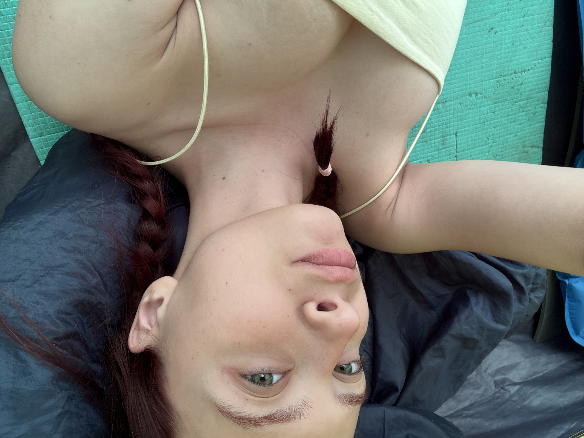 A woman with reddish-brown hair, styled in braids, is lying down, appearing to be inside a tent. She is wearing a light yellow tank top. The tent material is dark blue and gray, with a blue section visible in the background. A green mat is partially visible under her arm. The photograph appears to be taken from an overhead perspective. The image provides no information to suggest that the subject is in Khvalynsk, Russia.