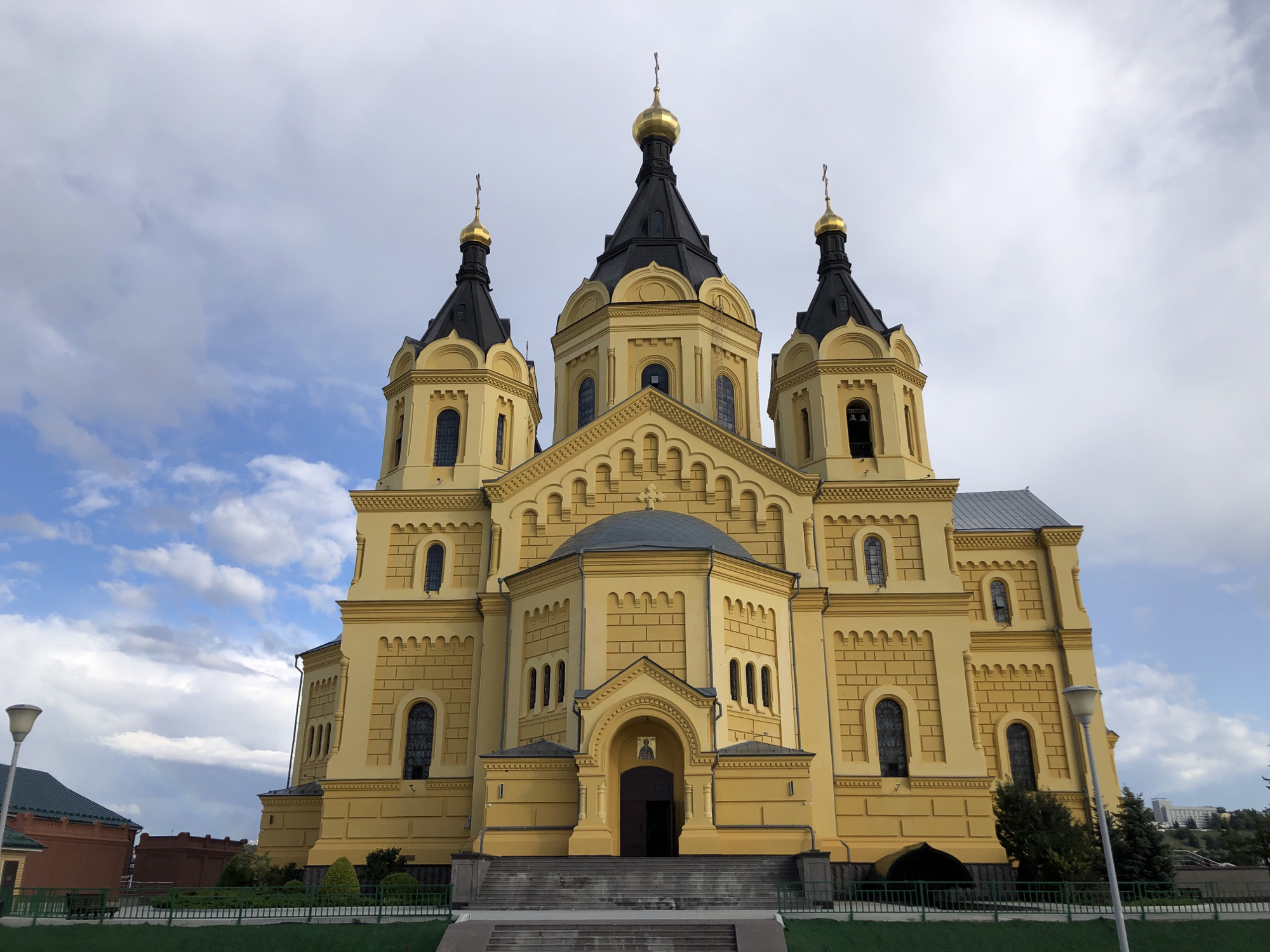 The image depicts the Alexander Nevsky Cathedral in Nizhny Novgorod, Russia, a prominent landmark in the city. The cathedral is a large, ornate building with a distinctive yellow facade and black onion domes topped with golden crosses. The main entrance is approached by a wide set of stone stairs. The sky above is a mix of blue and white clouds. In the foreground, a green fence runs along a manicured lawn. To the left of the cathedral, a red-brick building with a green roof and a modern street lamp are visible. In the background, trees and the outlines of distant buildings can be seen. The scene is captured from a frontal perspective, showcasing the grandeur of the cathedral. There are no people visible in the image.