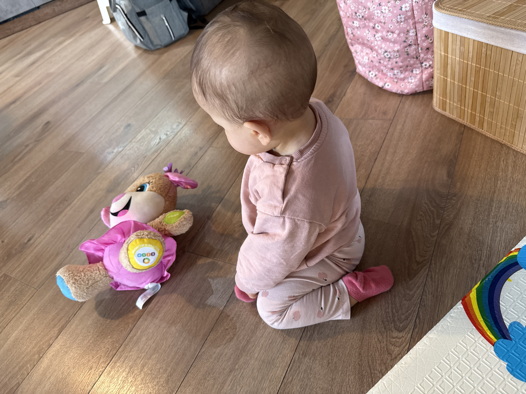 A baby is kneeling on a wooden floor next to a plush toy, in a room. The baby is facing the plush toy, with their head slightly turned to the left. The baby is wearing a pink long-sleeved shirt and patterned pants, and pink socks. The plush toy is brown and pink. A gray backpack is in the background, near the top left. A pink storage container and a bamboo box are in the background, near the top right. A white play mat with a rainbow design is in the lower right corner. The setting appears to be indoors. This location is most likely not Sofia, Bulgaria.