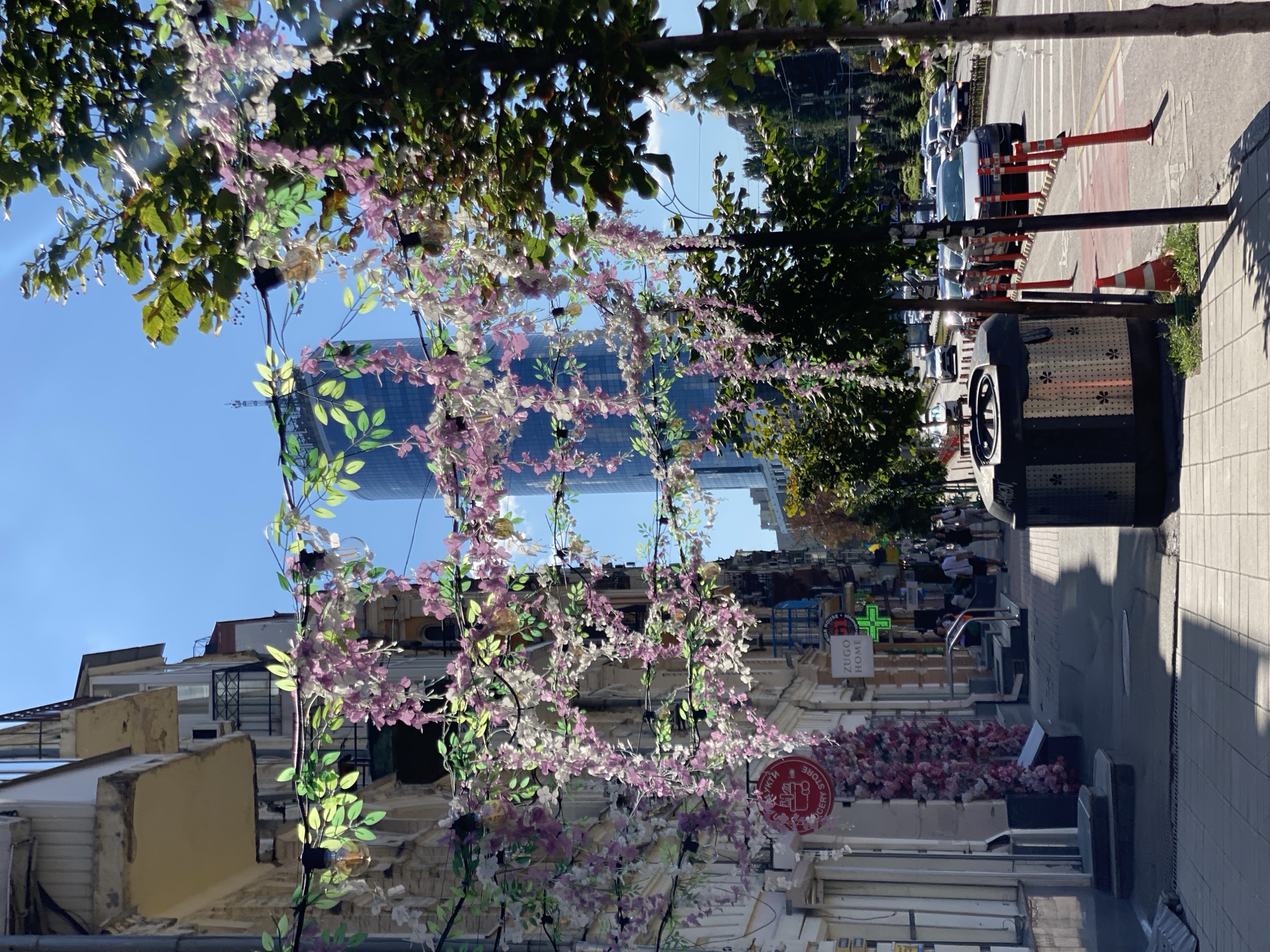 An outdoor, eye-level, wide shot captures a bright, sunny day in a city. The foreground is dominated by a pedestrian sidewalk lined with buildings on the left and trees on the right. Hanging decorations of artificial pink and white flowers and green leaves are strung across the street between the buildings. A modern, black trash receptacle with a decorative pattern is positioned on the sidewalk in the middle-ground. Beyond the sidewalk, a street with moderate traffic flows, marked by orange traffic cones and a pedestrian crosswalk. In the background, a tall, modern glass skyscraper stands against a clear blue sky, with older buildings visible between the skyscraper and the foreground. A few pedestrians are walking on the sidewalk, and several cars are driving on the street. The scene appears to be a street decorated for a festive occasion, possibly a market or a special event.