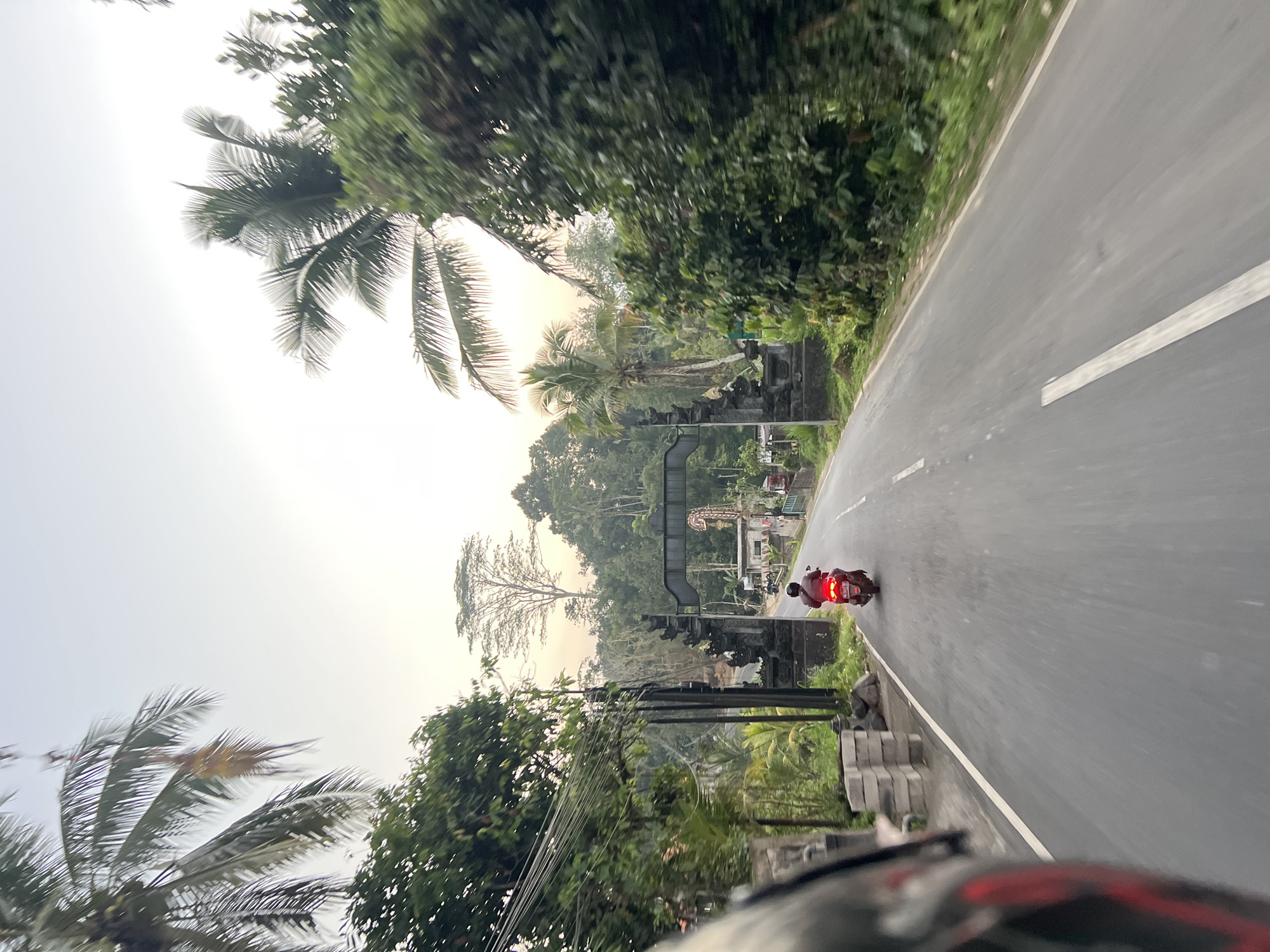 In Tembuku, Indonesia, an individual is riding a red scooter on an asphalt road. The scooter is moving away from the viewer, its brake lights illuminated. On either side of the road, lush green trees and palm trees line the path. In the distance, a traditional Balinese gate structure is visible, with intricate stone carvings. The sky is overcast with a warm, hazy glow, suggesting either dawn or dusk. The foreground of the image is partially obscured by a helmet, indicating the perspective is from another rider on a motorcycle.