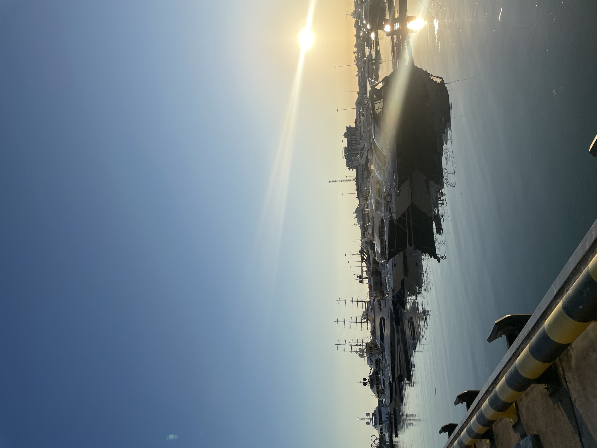 The image depicts a marina in Sochi, Russia, with numerous yachts and boats docked in the water. The sun is setting, casting a warm glow and creating reflections on the calm sea. The sky is a clear blue, with a slight haze near the horizon. The marina appears to be well-maintained, with a promenade visible in the background. There are no people visible in the image. The overall scene is peaceful and serene.