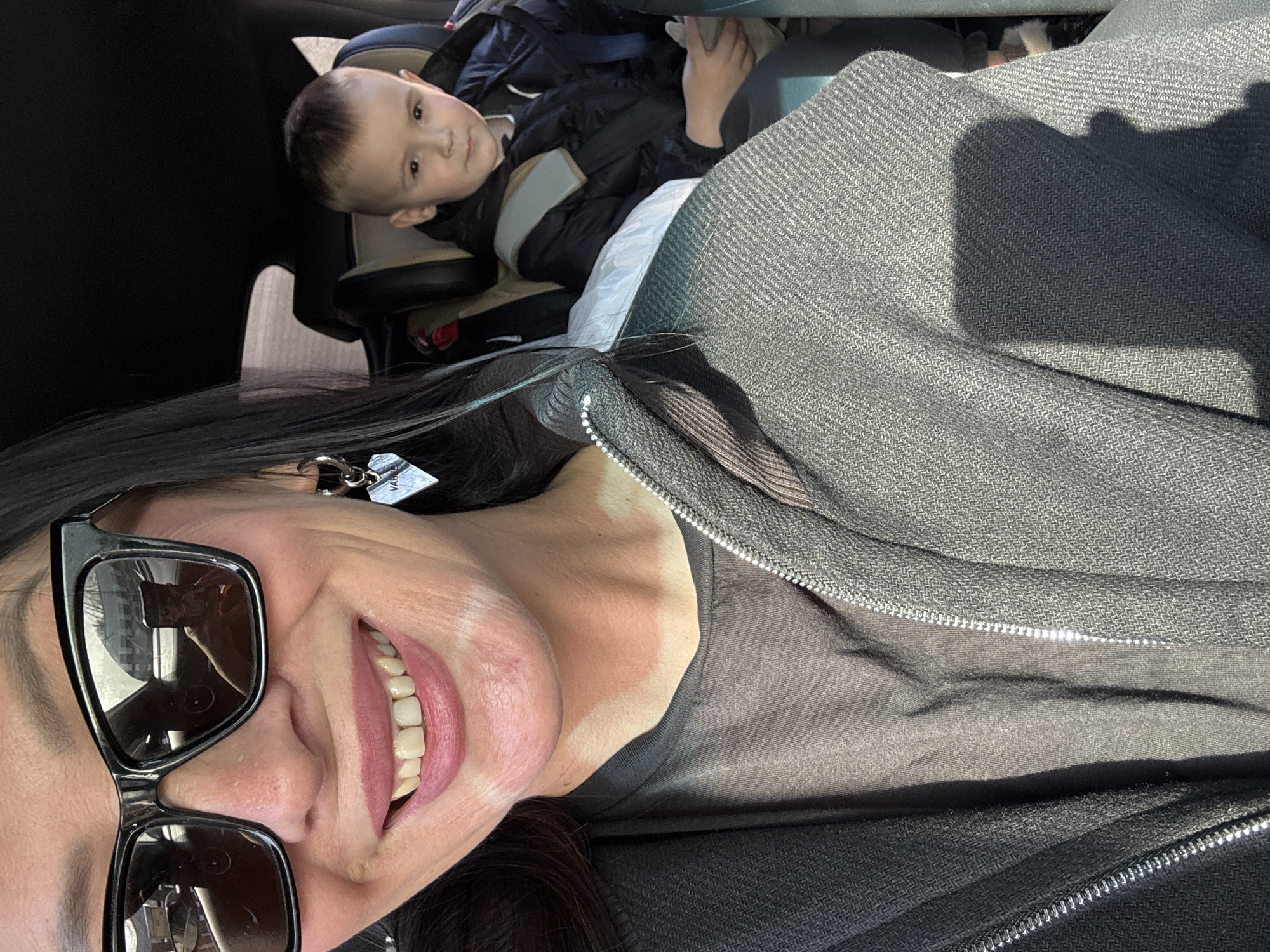 The image depicts two individuals inside a vehicle in Chaikovsky, Russia, illuminated by bright daylight. In the foreground, a woman with dark hair wears black sunglasses, a dark gray zippered jacket with a herringbone pattern over a dark t-shirt, and a silver-toned hoop earring with a heart-shaped charm on her left ear. She is smiling, revealing pink lipstick and areas of hypopigmentation on her chin and neck. The sunglasses reflect an outdoor scene featuring a road, trees, and a building, along with the smartphone used to capture the image. In the background, a young boy with short, light hair is seated in a beige and brown car seat. He wears a black padded jacket with light gray shoulder straps and looks directly towards the camera. A white plastic bag is positioned between the woman and the boy.