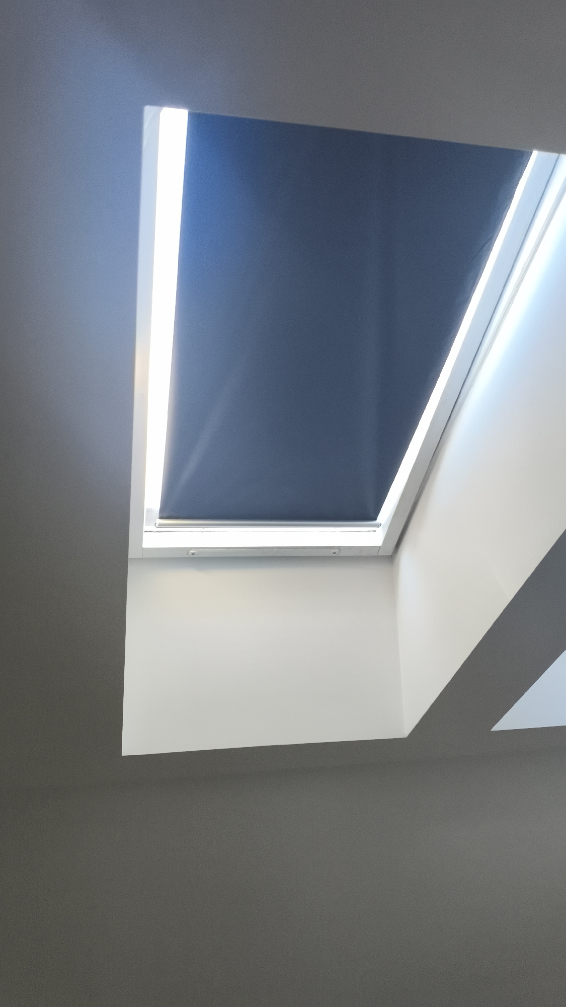The image shows a skylight in a room, seemingly indoors. The skylight is rectangular with a white frame and a dark blue roller blind that is partially closed. Light is filtering through the sides of the skylight, illuminating the frame. The walls surrounding the skylight are painted white, with a darker grey wall visible in the foreground. The location is unknown, but the user has specified that this is located in Nowy Tomyśl, Poland.