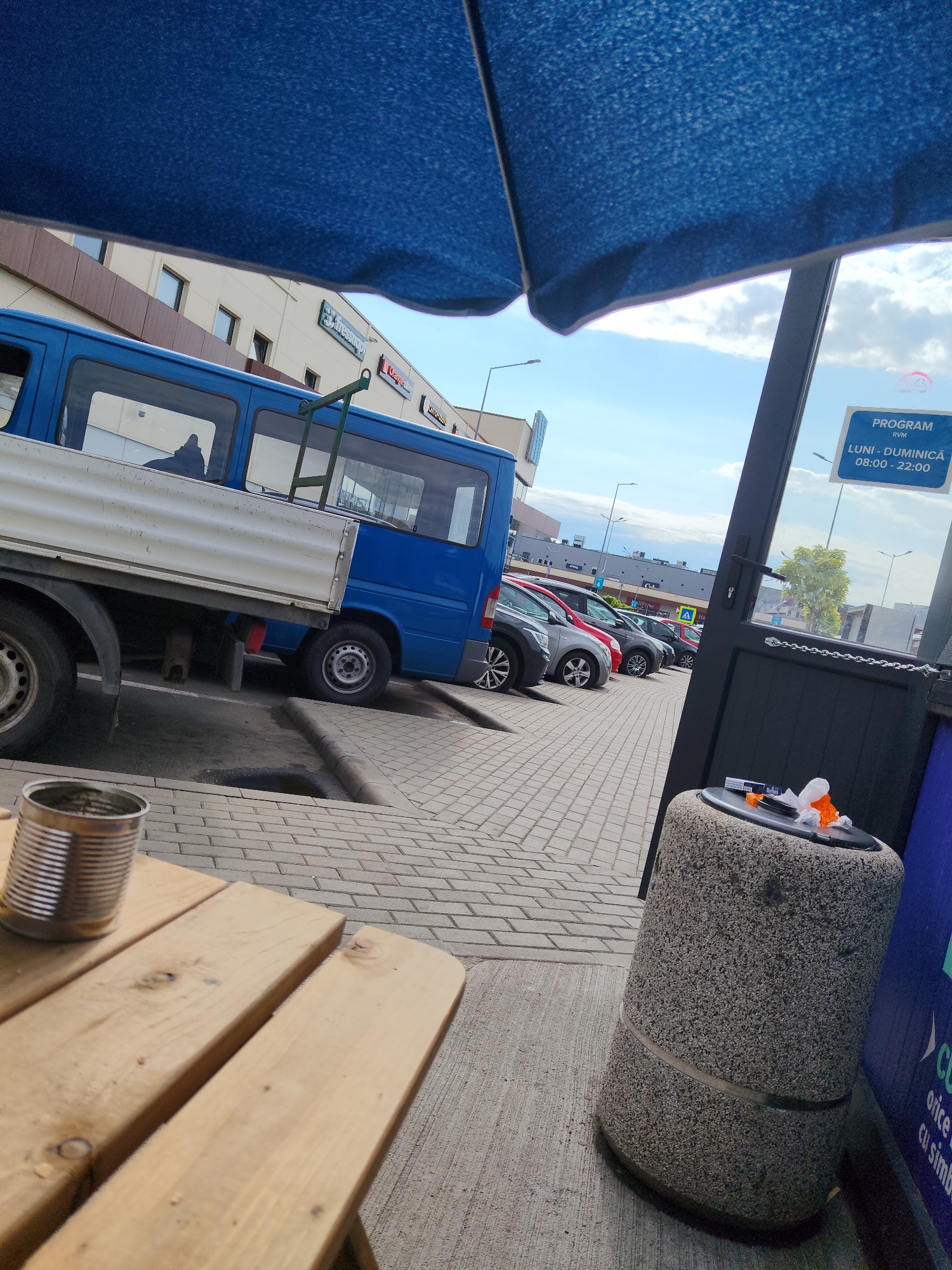 An outdoor scene in Arad, Romania, features a blue camper van parked next to a building. In the foreground, a metal can sits on a wooden table. A large blue umbrella partially obscures the upper portion of the image. In the background, several cars are parked in a lot. A sign on a dark door indicates opening hours from 08:00 to 22:00, Monday through Sunday. A stone trash receptacle with some orange and white refuse on top is situated next to the door.
