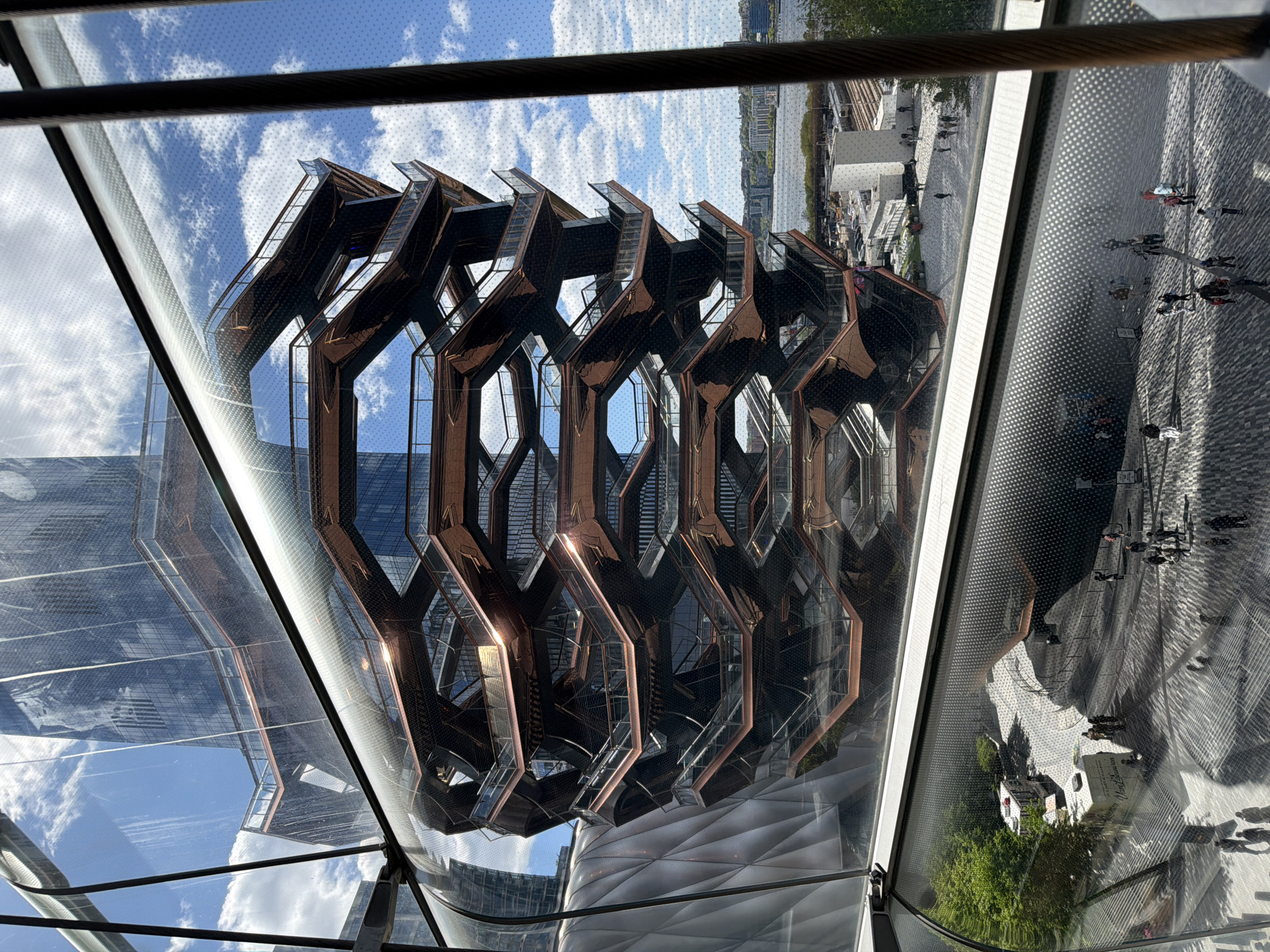 The image captures a view of the Vessel sculpture in Hudson Yards, New York City, as seen through a reflective surface, likely a window. The sculpture appears as a honeycomb-like structure of interconnected staircases and platforms, made of copper-colored steel. The reflection also reveals glimpses of the surrounding plaza, including paving stones, trees, and pedestrians. In the background, the Hudson River and the New Jersey skyline are visible. The sky is blue with scattered clouds. The reflective surface creates multiple layers of imagery, blending the Vessel with its surroundings and generating a distorted, artistic composition.