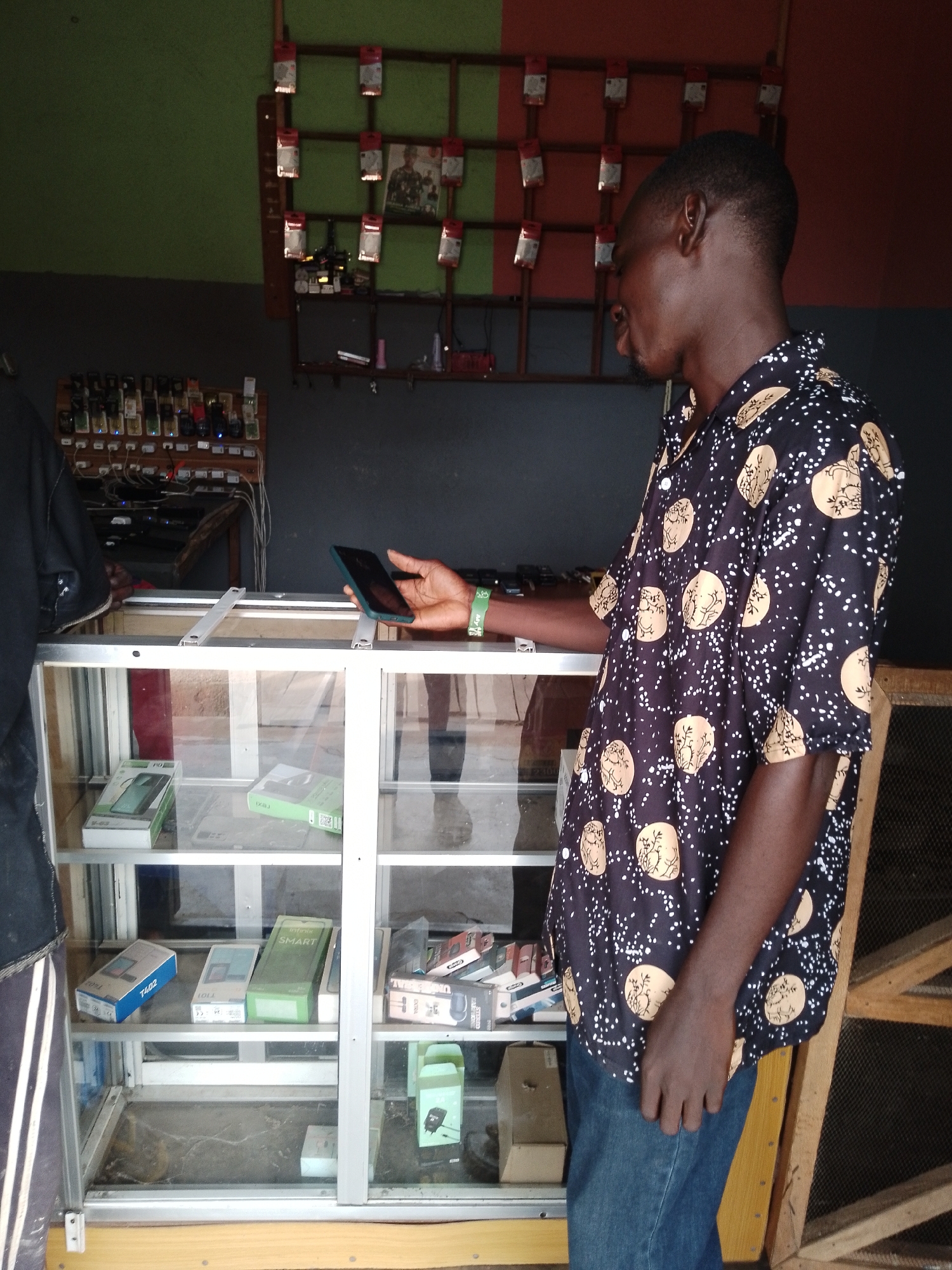 In a mobile phone shop in the city of Balewa, Nigeria, a young man in a black shirt with a patterned design stands by a glass display case. The shop has green and red walls. The display case is filled with various mobile phones and accessories. On a shelf above the case, there are more mobile phones displayed on a wooden board. The man is holding a dark-colored smartphone in his right hand. He is looking down at the phone. The image appears to be taken indoors, possibly during the daytime.