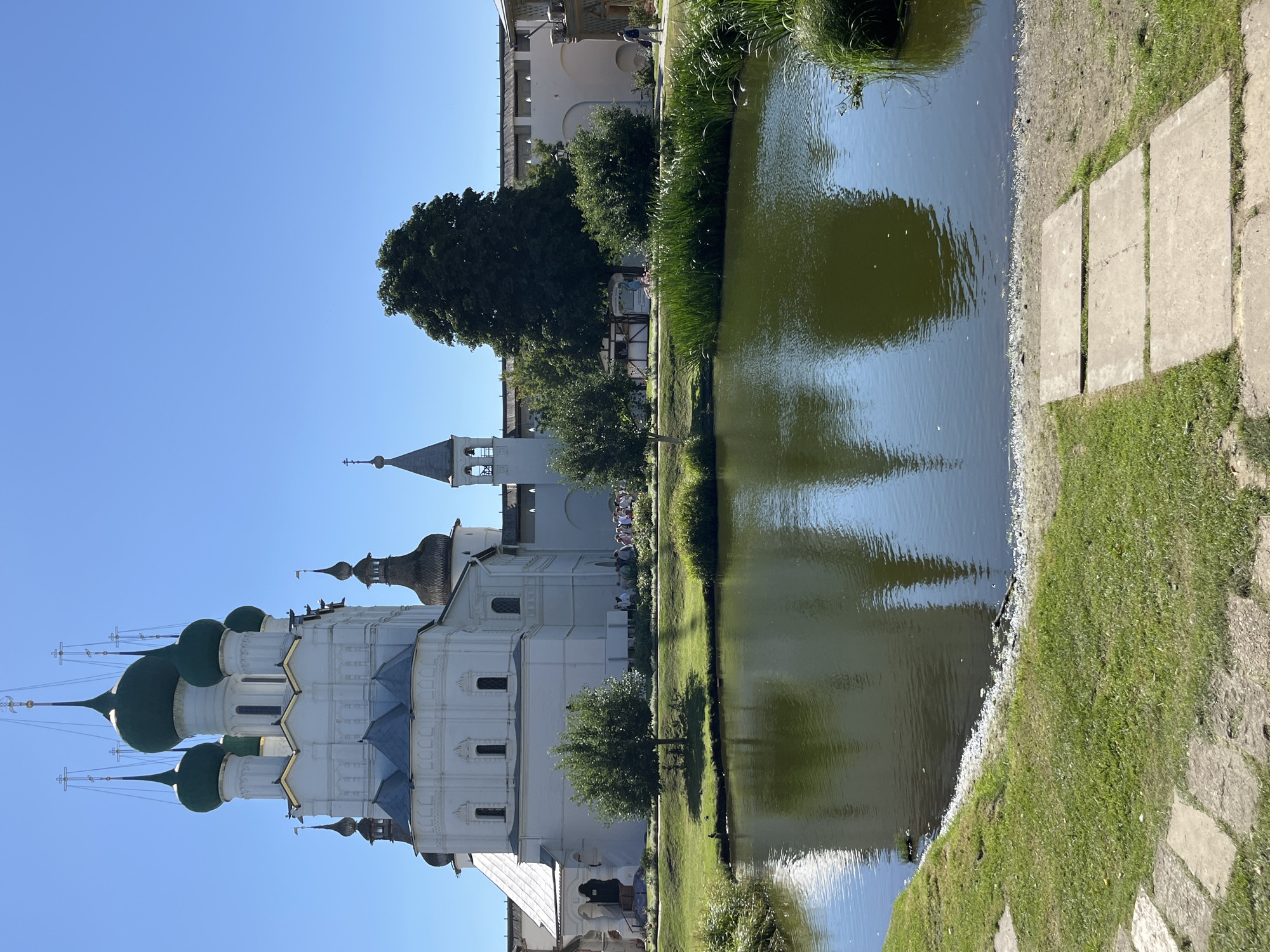 The image depicts the Rostov Kremlin in the city of Rostov, Russia, on a clear, sunny day. In the foreground, a calm body of water reflects the sky and surrounding architecture. The water is edged by lush green grass and a path made of flat, rectangular stones. Behind the water, a large, white building with multiple green domes and intricate architectural details stands prominently. This building appears to be a church or cathedral, with tall spires and crosses. To the right of the main structure, a white wall with a wooden crenelated top, characteristic of a fortress or Kremlin, stretches into the background. Several trees, varying in size and foliage, are interspersed around the grounds, adding to the scenic view. A few people are visible in the middle ground, walking along the paths or gathered near the building, suggesting it is a public or tourist site. The sky is a clear, bright blue, with no clouds present.