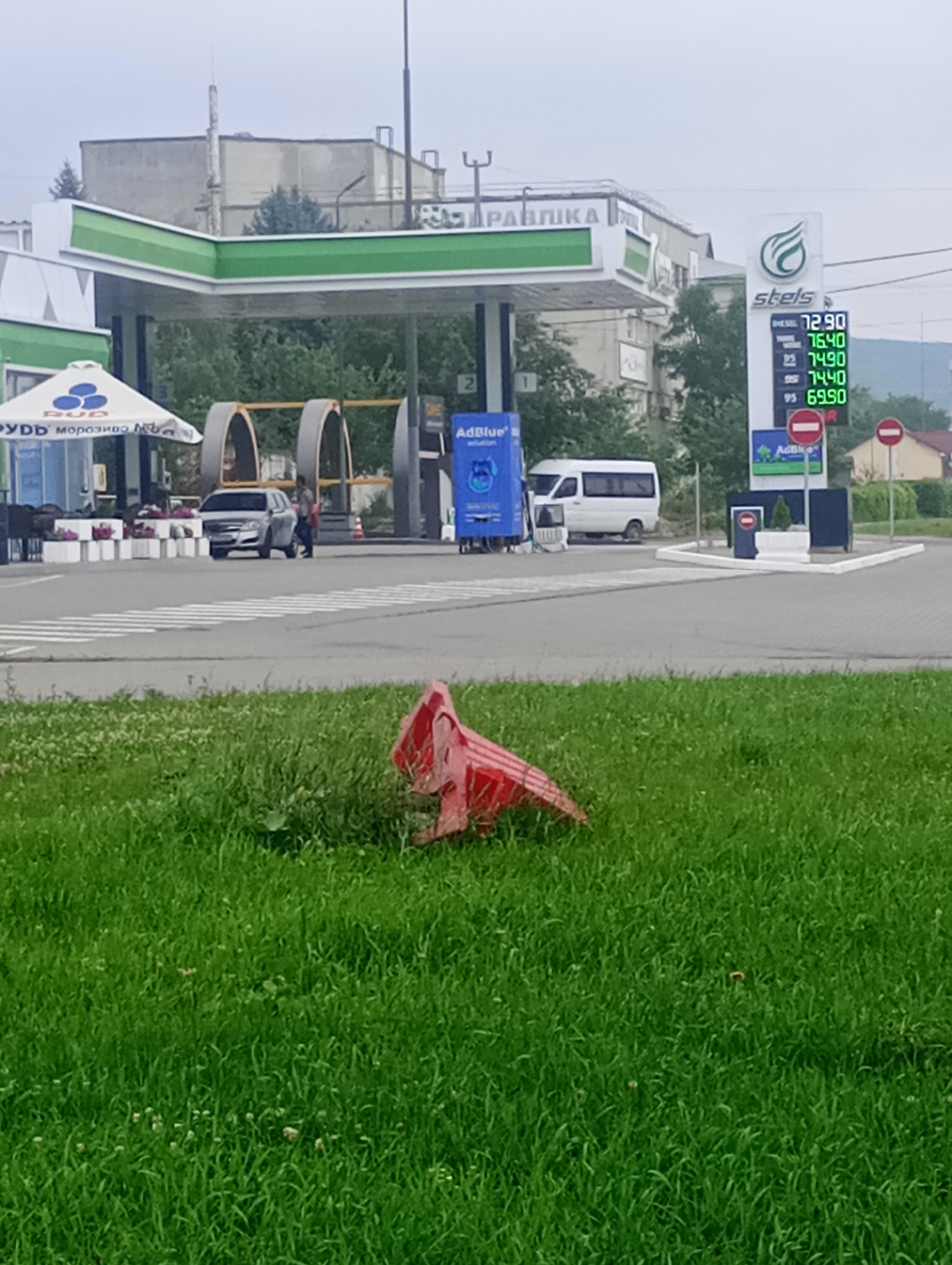 This media file depicts a gas station with a prominent "stels" brand sign displaying fuel prices. The sign indicates prices for diesel at 72.90, 76.40, 74.90, and 74.40, and for 95 octane gasoline at 69.90. A blue dispenser labeled "AdBlue" is also visible. The gas station has a green and white canopy over the pumps. In the background, there are buildings, trees, and a hilly landscape. To the left of the gas station, an umbrella with the logo "RUD" and text "морозиво ва" is set up, suggesting a convenience store or cafe attached to the station. A silver car is parked at one of the pumps, and a person is walking nearby. A white van is also present. In the foreground, a red plastic barrier lies on a patch of green grass. The lighting suggests it is daytime, and the sky appears overcast. The scene appears to be an outdoor commercial setting in a somewhat rural or suburban area. The overall impression is a functional, everyday scene at a fueling station.