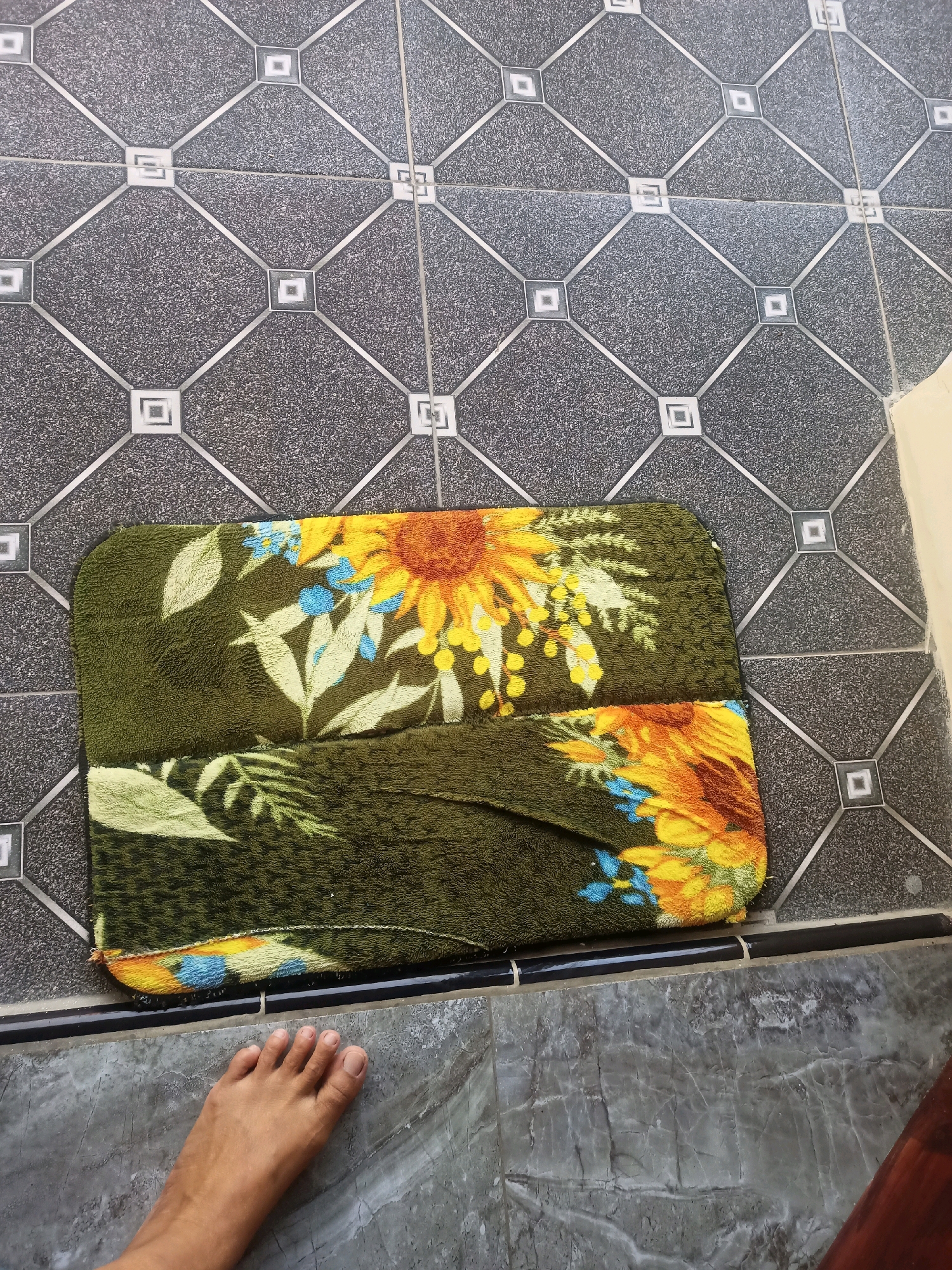 In Karangtalun, Indonesia, a rectangular bath mat with a floral pattern of sunflowers, blue flowers, and green leaves lies on a floor tiled with dark gray squares featuring a diamond pattern. The bath mat is positioned on top of the patterned tiles. In the lower left corner of the image, a bare human foot is visible, positioned on a different type of flooring that appears to be gray marble. The transition between the two flooring types is marked by a thin black strip. The scene depicts an indoor or semi-outdoor area, potentially a entryway or bathroom.