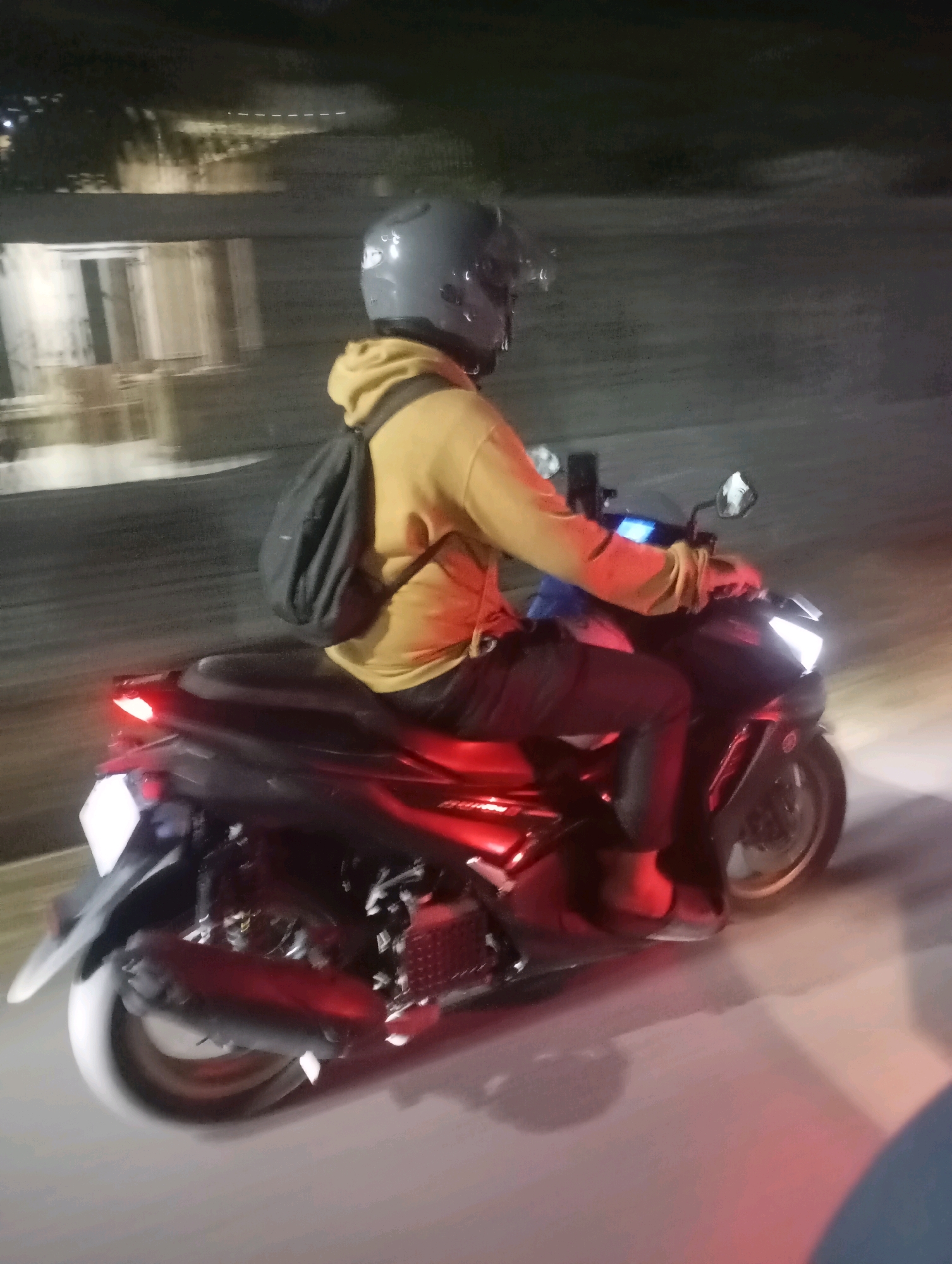 A person wearing a yellow hoodie and a gray helmet is riding a black and red motorcycle at night. The motorcycle has a license plate on the back. The scene appears to be a street in Babelan, Indonesia, as indicated by the blurred background that shows a building with lights and a road surface. The image is taken from a slightly elevated side angle, capturing the rider and the motorcycle in motion. The lighting suggests it is nighttime or very low light conditions, with the motorcycle's headlights and taillights illuminated. The overall image has a slightly motion-blurred quality, typical of a moving subject captured in low light.