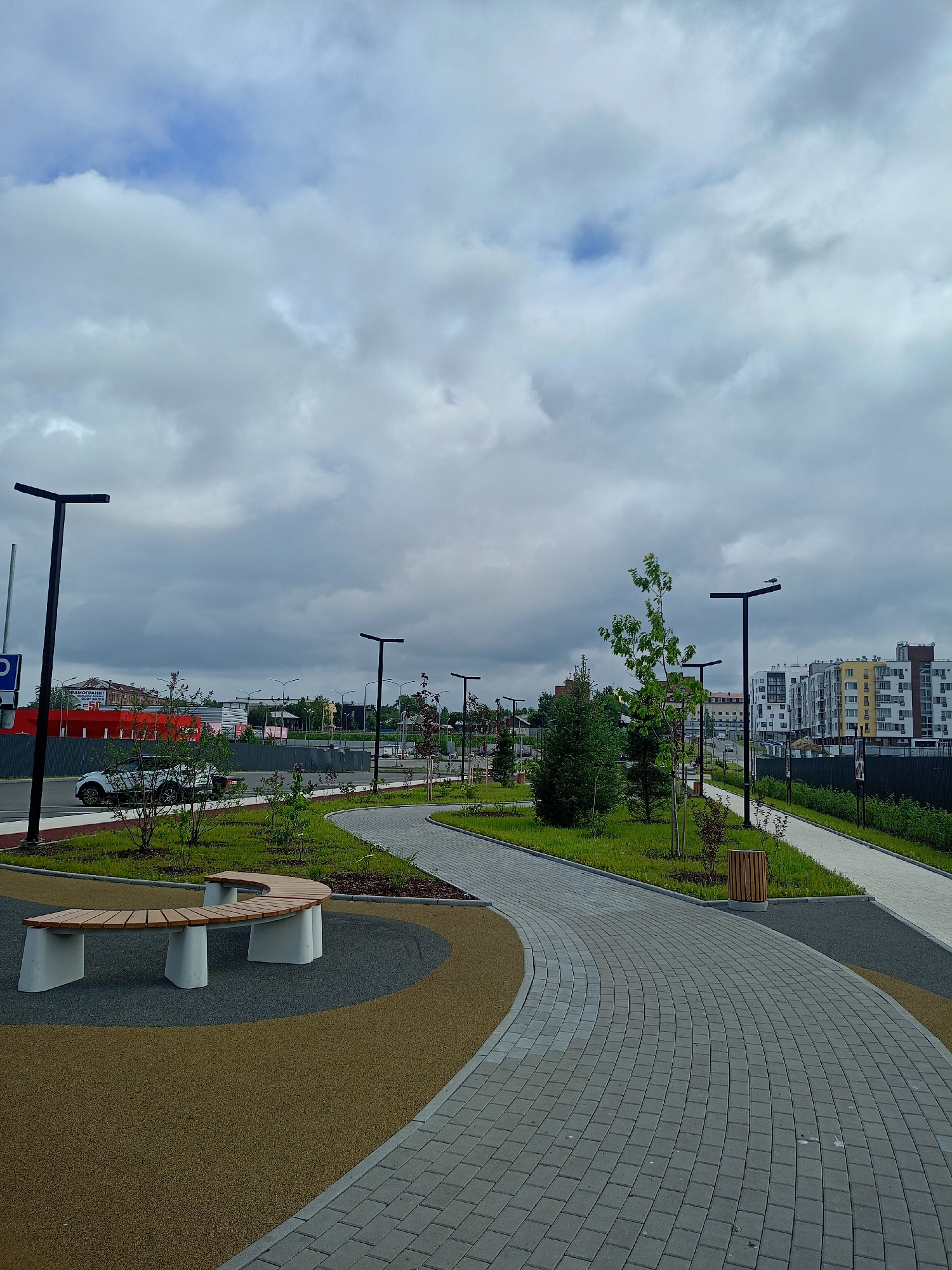 An outdoor scene depicts a public park or walkway with a paved path winding through it. The path is made of grey interlocking pavers and curves to the right in the foreground, then continues into the background. The walkway is bordered by grass, small trees, and shrubs. There is a curved wooden bench with concrete supports on the left side of the image, set on a circular area with a speckled yellow and grey surface.
In the background, several modern apartment buildings with light-colored facades and visible windows are situated behind a black fence. A row of black lampposts with modern rectangular light fixtures line the path. A white car is parked on a road to the left of the path, with more buildings and a red structure visible beyond. The sky is overcast with dramatic grey clouds interspersed with patches of blue. The overall impression is a newly developed urban green space under a cloudy sky.