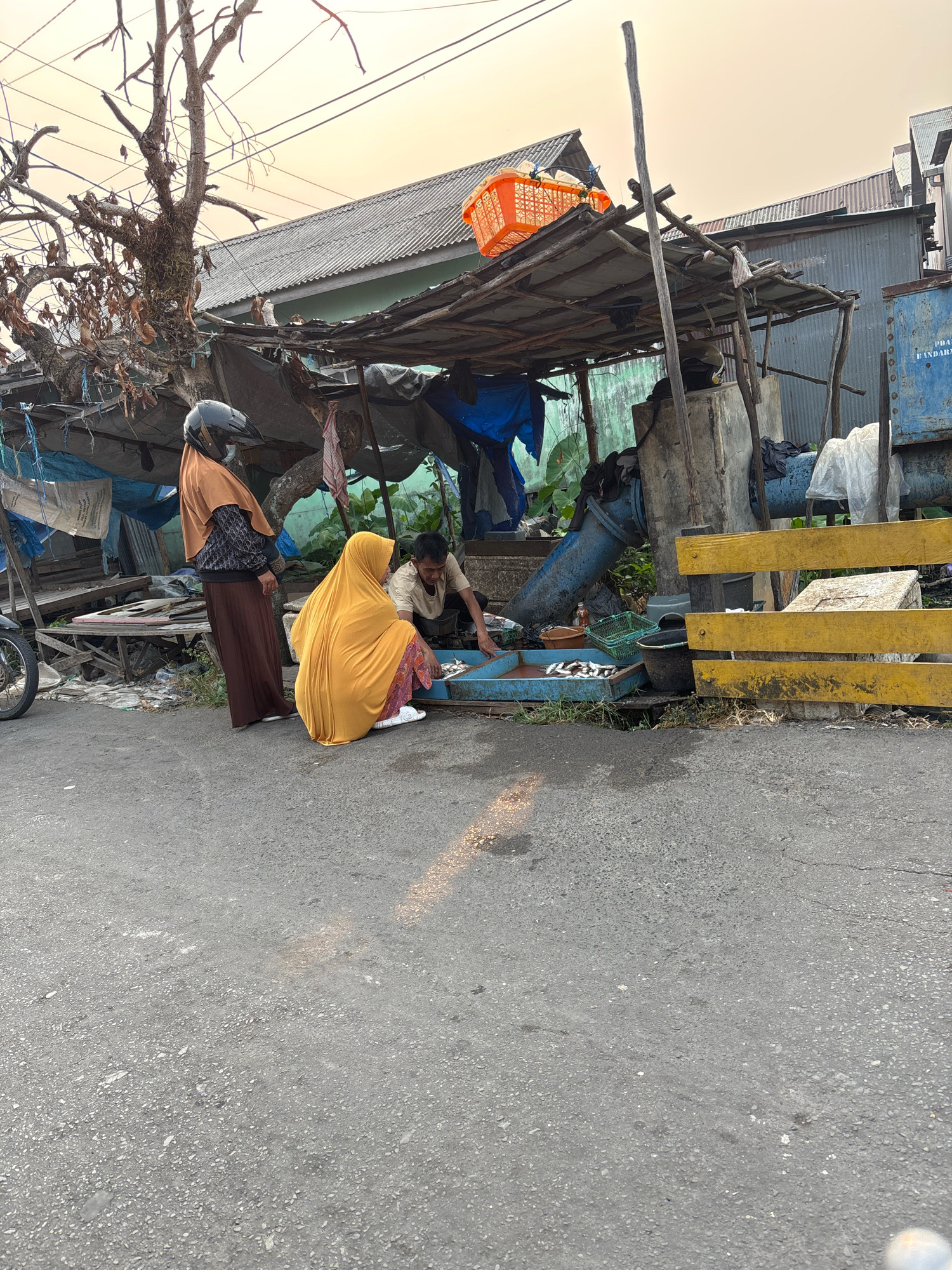 The image depicts an outdoor scene, likely a street market in Banjarmasin, Indonesia, given the context of selling fresh produce. The focal point is a makeshift stall constructed with wood and tarps, under which three individuals are interacting. A person wearing a yellow hijab and patterned skirt is kneeling, attending to a blue tray filled with small fish. To their left, a person wearing an orange hijab and a black and white patterned top stands, holding a black helmet. Another person, partially obscured, is crouched behind the tray, possibly assisting with the fish. The stall itself is cluttered with various items, including blue tarps, plastic containers, a green basket, and what appears to be large blue pipes in the background. Behind the stall, a corrugated metal building and parts of other structures are visible, suggesting an urban or semi-urban environment. The ground is paved asphalt, and a dried-out tree with sparse branches stands to the left of the stall. The sky is a pale orange, indicating either dawn or dusk.