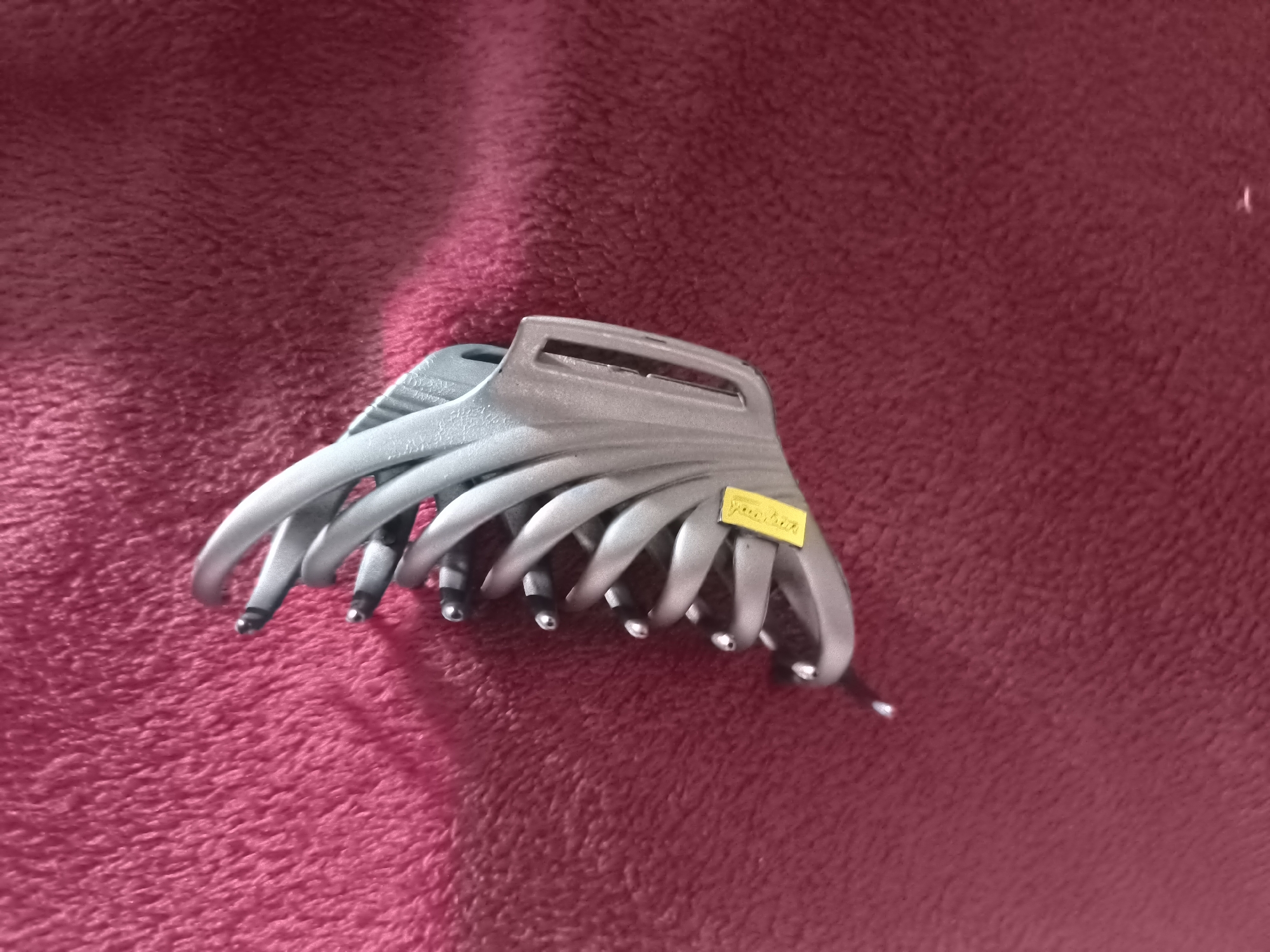 An overhead shot shows a light gray hair claw clip on a red fuzzy surface. The clip has several interlocking teeth that curve upward and inward. A small yellow sticker with black text is adhered to the top of the clip. The surface has a soft texture and is lit from the left, casting a shadow from the clip onto the surface.
