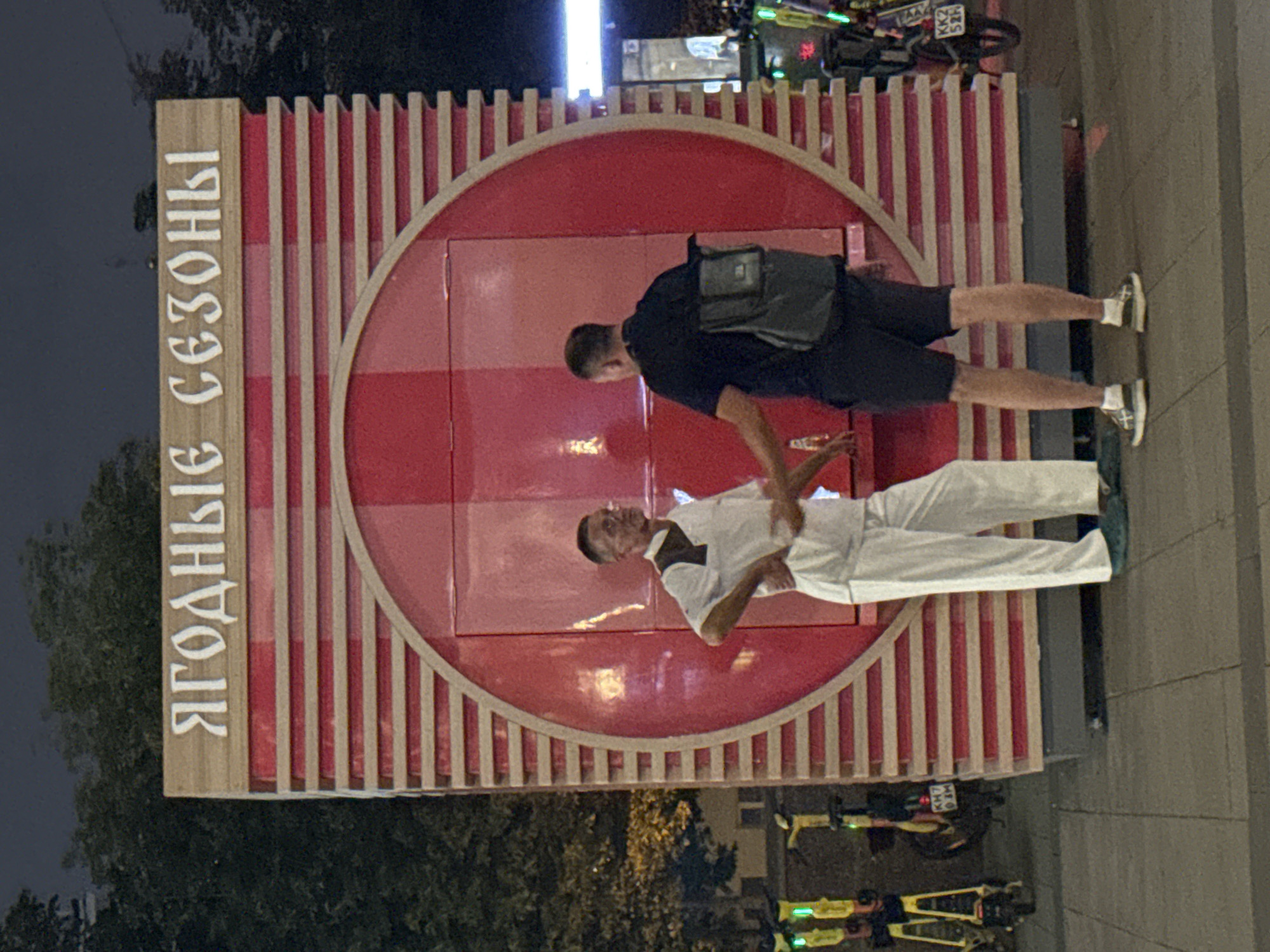 In Moscow, Russia, two men are standing in front of a red, circular kiosk with wooden slatted siding. The kiosk has Cyrillic lettering on top that reads "ЯГОДНЫЕ СЕЗОНЫ" (Berry Seasons). The man on the left is wearing a white short-sleeved shirt with a dark collar and white pants, and he has his left hand on his hip. The man on the right is wearing a black t-shirt, dark shorts, and a black backpack, and he is shaking the other man's hand. The ground is paved with grey concrete tiles. Several yellow electric scooters are parked to the left of the kiosk. The scene appears to be outdoors in a public space, possibly a street or plaza, at night, with trees visible in the background and artificial lighting.
