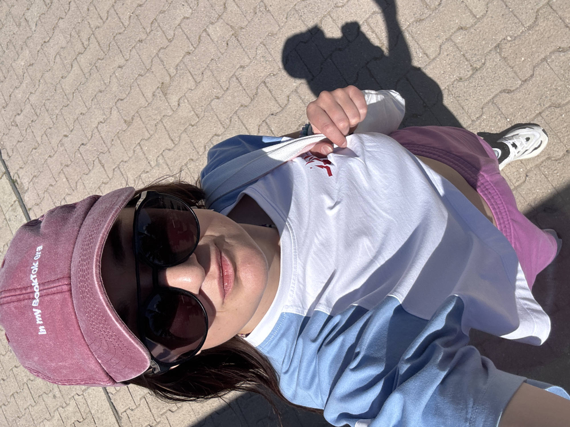 A woman lies on a paved surface, likely a sidewalk or plaza. She wears sunglasses, a pink cap embroidered with "In my BookTok era", a white t-shirt with blue sleeves, and pink pants. One hand holds the fabric of her t-shirt. Part of a white and black shoe is visible near her leg. The location is an outdoor area within the city of Unknown, Unknown. The time of day is daytime. A human shadow is seen on the paved surface near the upper right corner of the image.