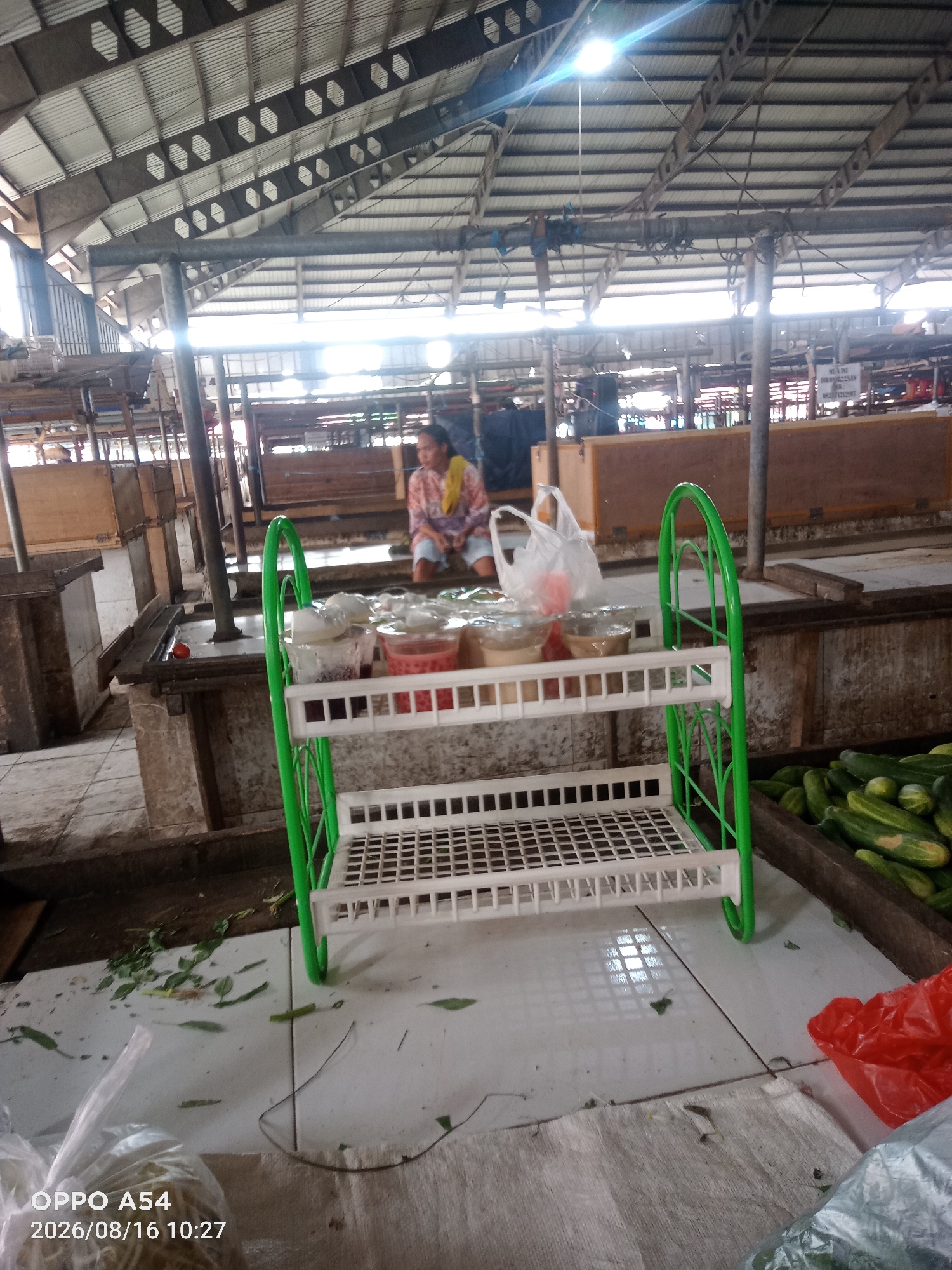 The image depicts an indoor market scene in Jambi, Indonesia. A person is seated in the background, and in the foreground, a white plastic shelf with a green frame holds several transparent containers of food items. To the right of the shelf, a wooden crate contains a pile of cucumbers. The market appears to be an open-air structure with a corrugated metal roof supported by metal beams and pillars. The floor is tiled, and there are scattered green leaves on the ground, indicating ongoing market activity.