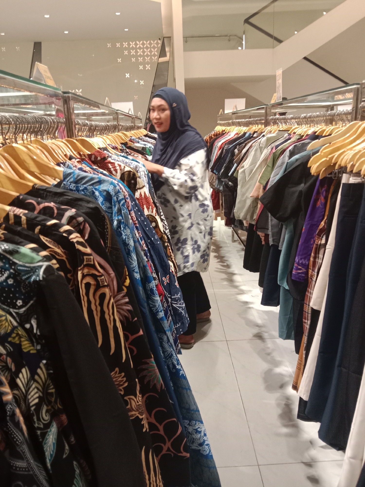 A woman wearing a dark blue hijab and a patterned tunic is browsing clothes in a store. She is standing to the right of a rack of colorful batik garments, with her hand reaching towards them. The store appears to be well-lit with fluorescent lighting. The background shows more clothing racks, mirrors, and decorative patterns on the wall. The floor is tiled with a light color and shows some reflections. This scene is likely in a department store or clothing boutique located in Semarang, Indonesia.