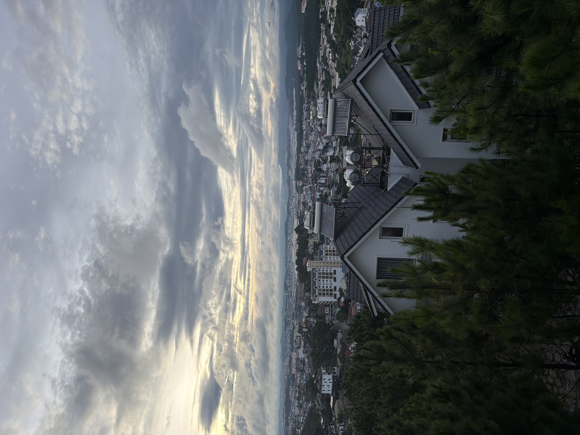 The image captures a panoramic view of Xuân Trường ward in Đà Lạt, Vietnam, under a dramatic, cloudy sky. In the foreground, two modern white houses with dark gray pitched roofs are partially obscured by lush, dark green pine trees. The roofs of these houses are equipped with solar water heaters.
Beyond the houses, the city of Đà Lạt unfolds with a dense collection of buildings featuring various architectural styles, including white apartment blocks with visible windows and balconies, and other residential structures. The city stretches towards the horizon, where rolling hills and mountains, softened by a layer of atmospheric haze, form the backdrop. The sky is filled with a complex pattern of gray and white clouds, with streaks of pale yellow light suggesting either early morning or late afternoon sun breaking through. The overall impression is one of a sprawling urban landscape nestled within a verdant, mountainous region, under a moody and dynamic sky. There are no people visible in the frame.