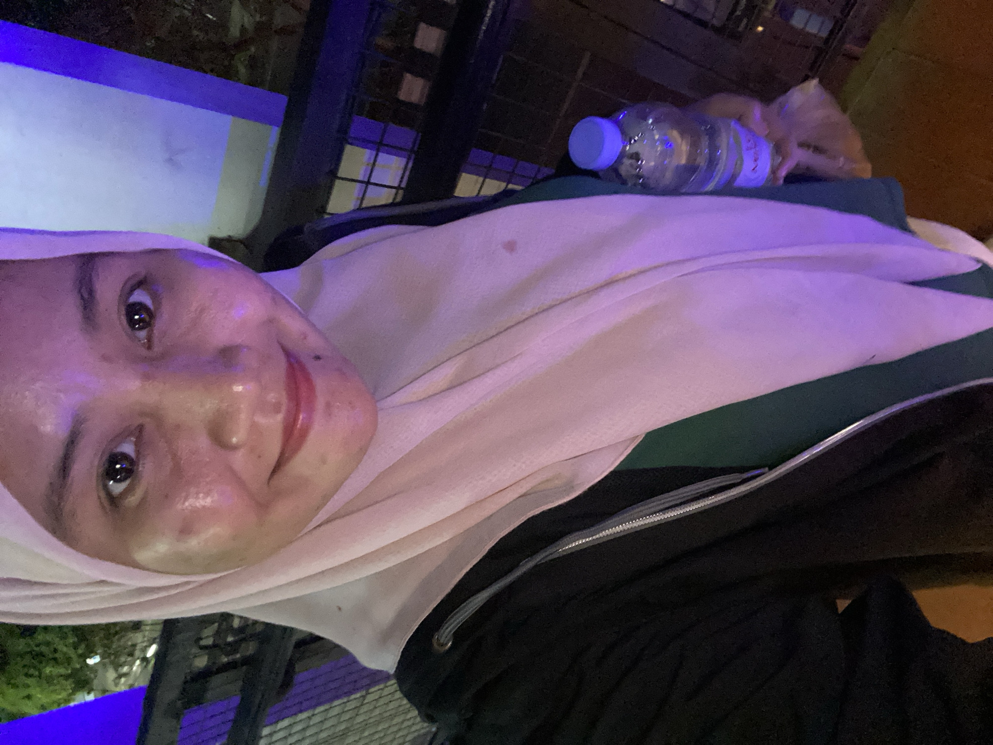 In this selfie, a person wearing a light pink hijab and a black jacket is holding a clear plastic bottle of water in their right hand. The person's face is partially visible, with their eyes looking at the camera and their mouth curved in a slight smile. The background is out of focus but appears to be an outdoor setting with purple lighting illuminating a dark railing and some greenery. The image was taken in Banjarbaru, Indonesia, likely in a public area or a venue with decorative lighting.