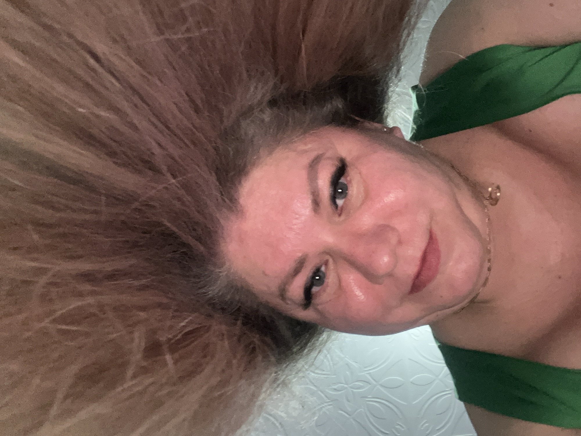 A woman with long, light brown hair is lying down, looking at the camera. She is wearing a green top. A gold necklace is visible around her neck. The background appears to be a white surface with a repeating pattern. There is no evidence to indicate the location is Izhevsk, Russia.