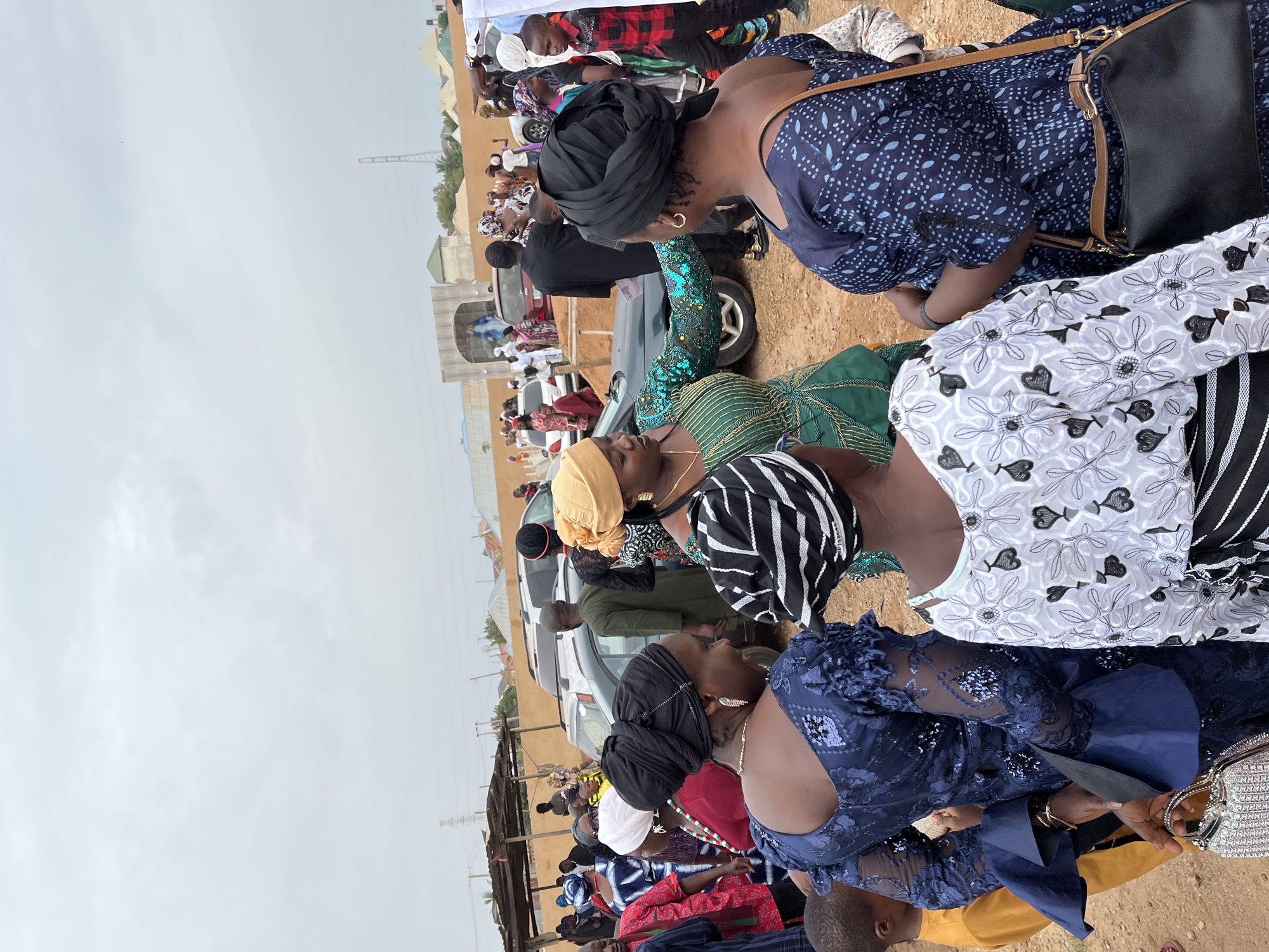 A gathering of people in Jos, Nigeria, is visible in the image. The focus is on a group of women seated together, dressed in various colorful and patterned traditional African clothing and headwraps. Cars are parked behind them, suggesting the event may be outdoors near a road or parking area. Other people are seen standing or sitting in the background, indicating a larger gathering. The setting appears to be a dry, dusty area with simple buildings and utility poles in the distance. The sky is overcast, suggesting the time of day.