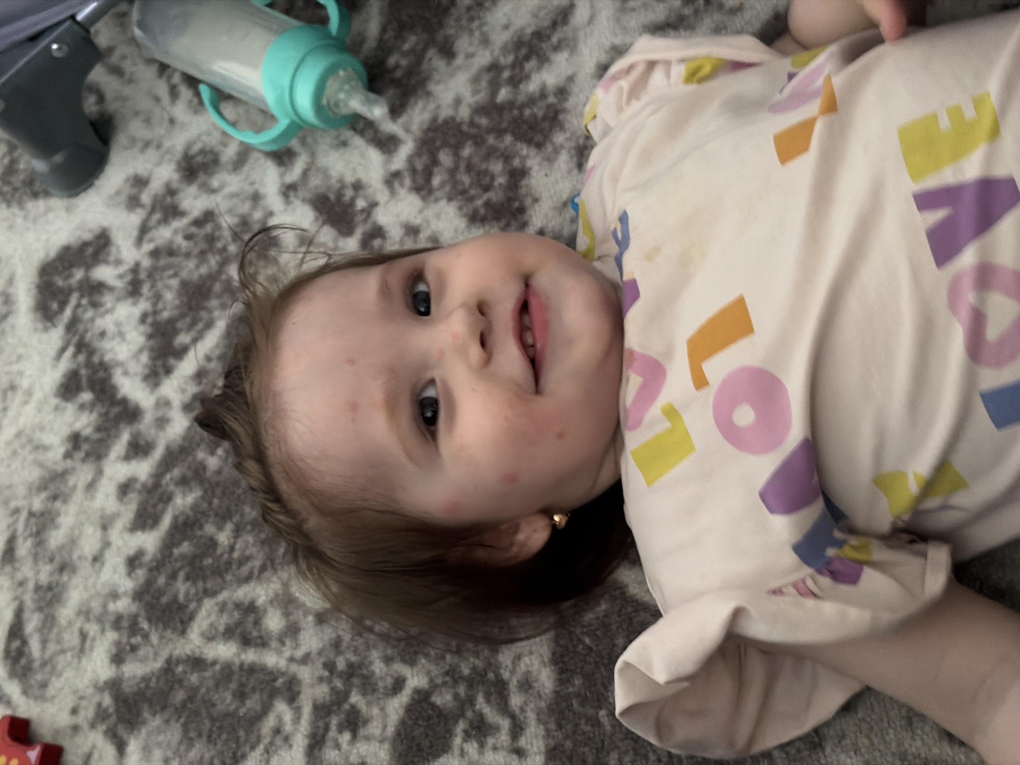 A young toddler with fair skin and brown hair lies on a grey and white speckled rug. The toddler is wearing a pale pink t-shirt with colorful letters that spell out "LOVE" in orange, yellow, purple, and pink. The toddler has a small gold heart-shaped earring in their left ear. There are red spots on the toddler's cheeks and forehead. In the background, a baby bottle with a green nipple and handles is visible, along with a part of a grey object which appears to be a baby walker or stroller. The lighting suggests an indoor setting. There is no visual information to determine the location within the city of Chaikovsky, Russia.