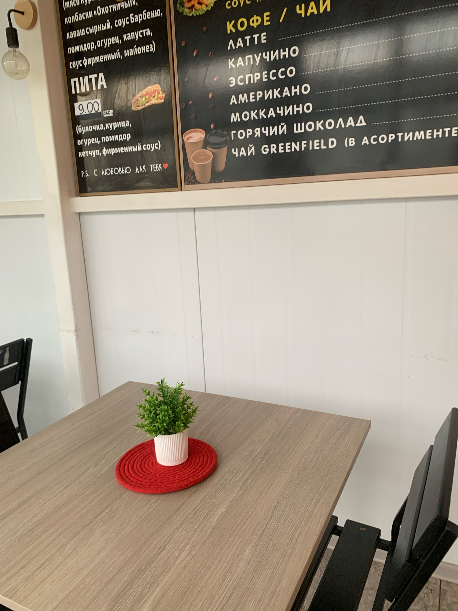 The image shows a close-up of a menu board in a cafe in Kapyl, Belarus. The menu board features various coffee and tea options, including latte, cappuccino, espresso, americano, mokkachino, hot chocolate, and Greenfield tea. It also lists a "Pita" dish for 9.00, with ingredients described as bun, chicken, cucumber, tomato, ketchup, and special sauce. The menu is written in Russian. The cafe environment appears casual, with wooden tables and chairs, and a small potted plant on one of the tables. A light fixture is visible on the left side of the image.
